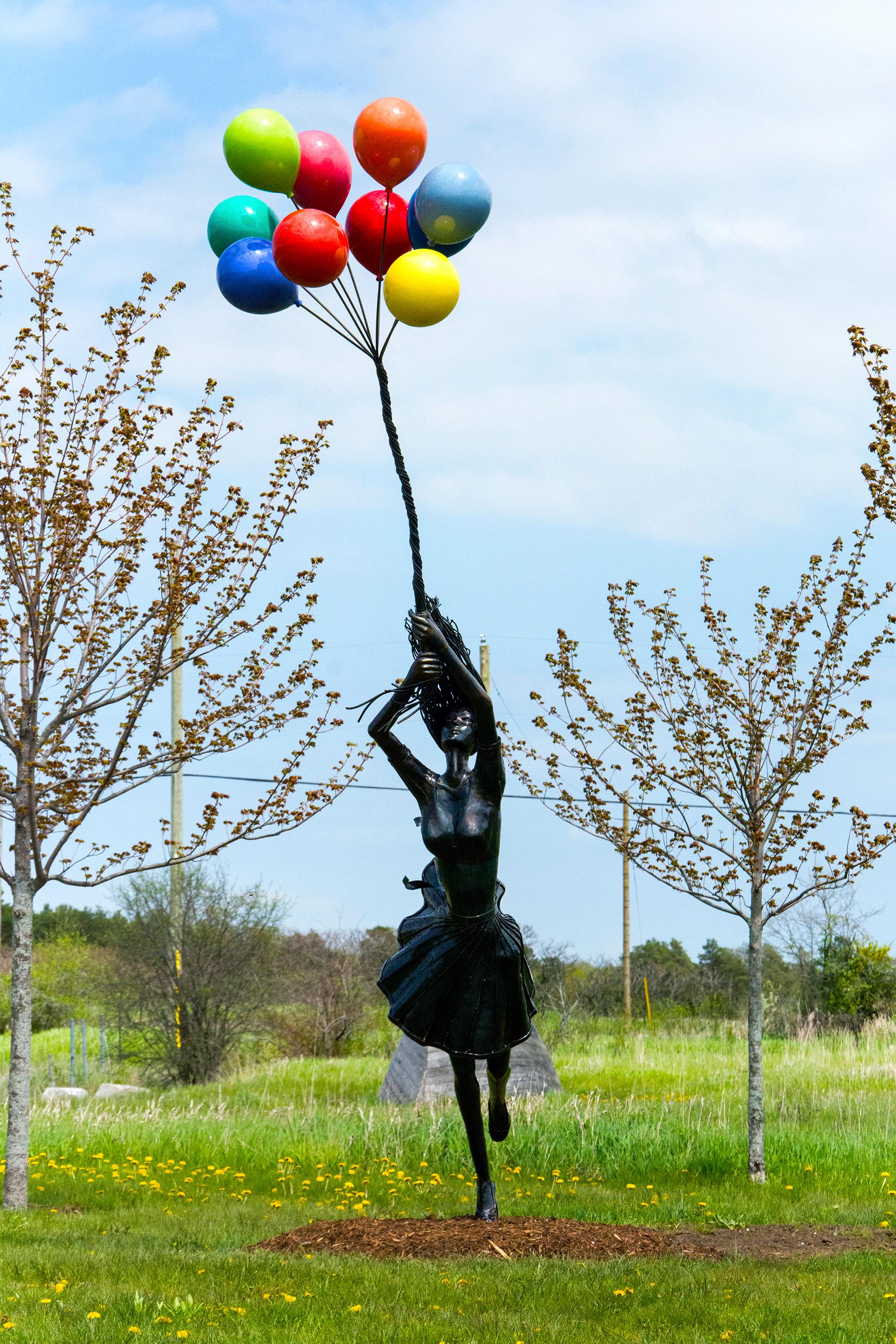 Rendered in fine detail, the figure of a young woman is carried upwards by colorful balloons in this provocative outdoor sculpture. Derya Ozparlak's sculpture bring various character types and identities  to life. Her sculptures portray the