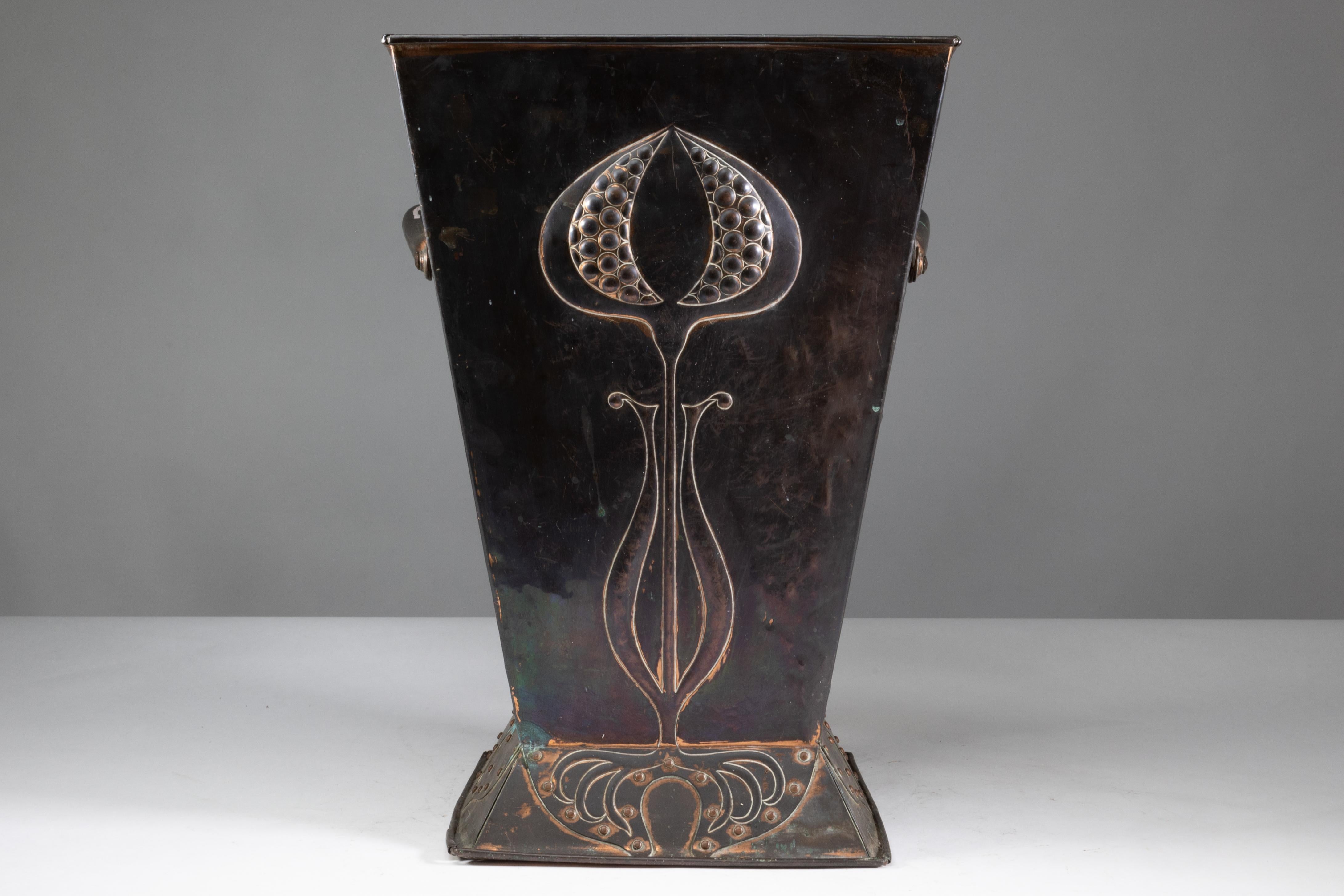 Robert Hilton for Hilton Ware. 
An impressive Arts and Crafts copper coal bucket with a single flowering seed pod, the roots of which are of blind riveted fretwork to the base. The sides with similar riveted decoration to the base, flowing up with
