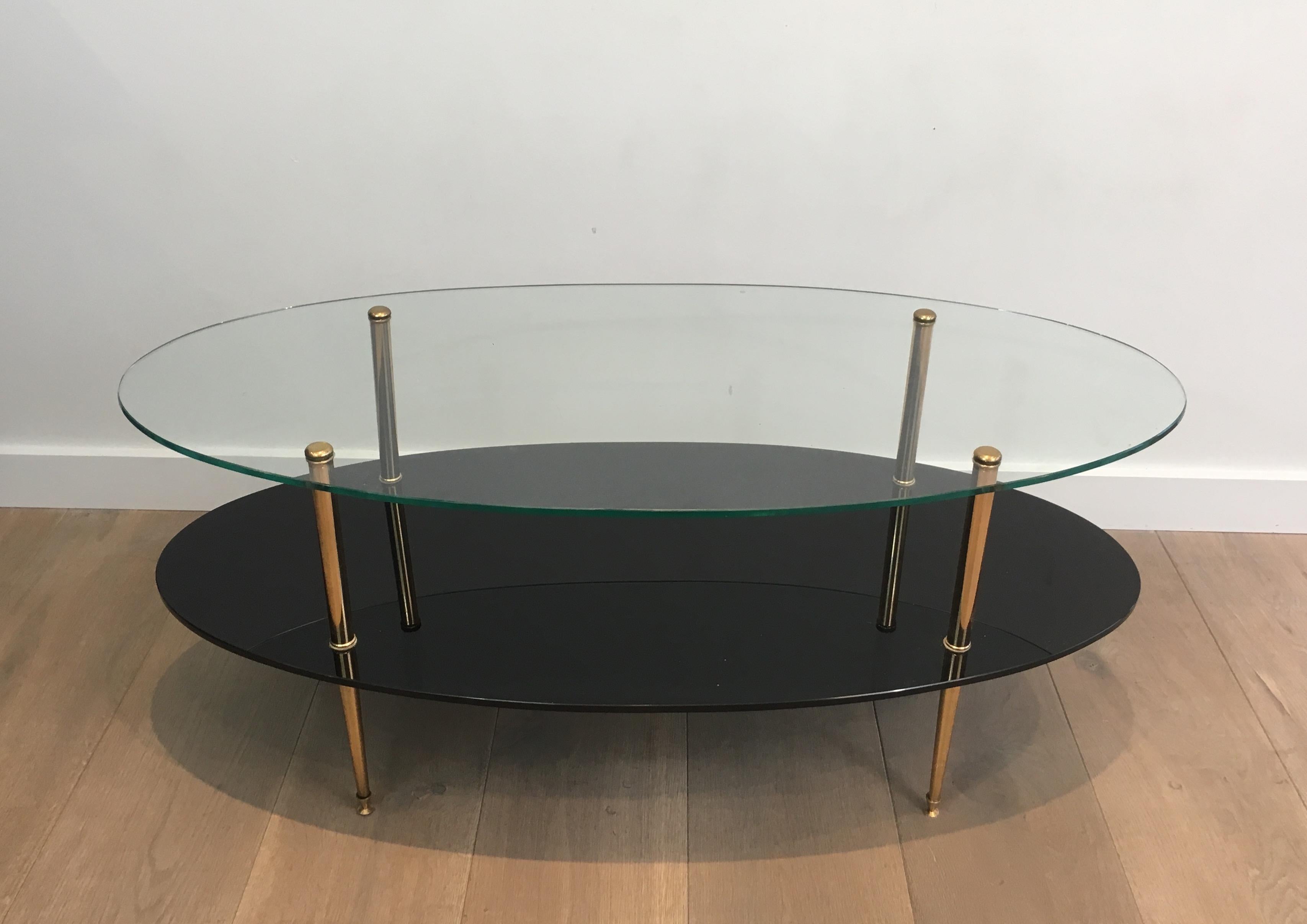 This design oval coffee table has a clear glass as top shelf and a black lacquered glass as bottom shelf. The feet are made of brass and both shelves are linked by these simple brass feet on top of which is a round brass finial. This cocktail table