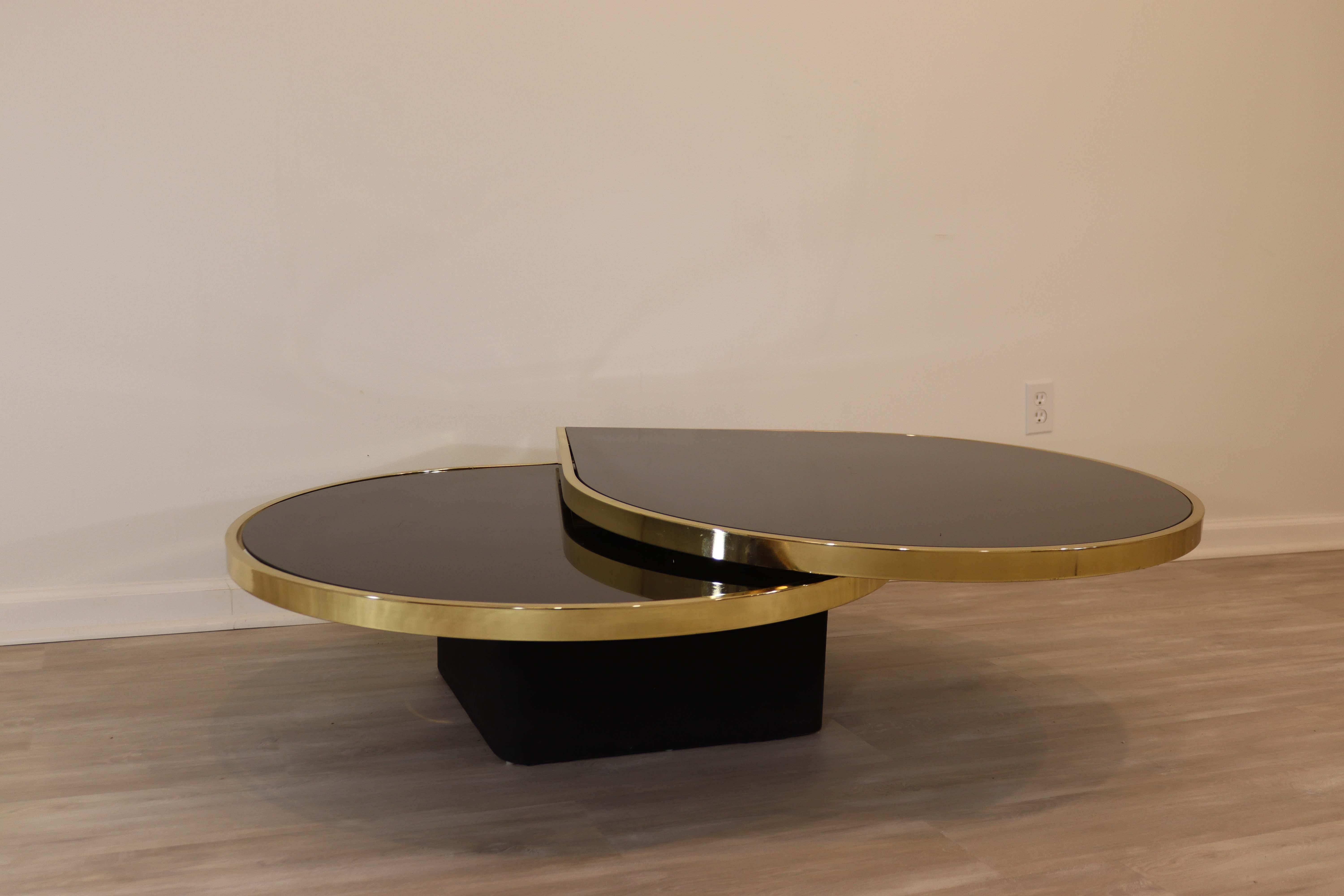 Up for sale is this modern 2-tier coffee table in black glass and brass details from the Design Institute of America circa the 1970's. Each tear drop swivels independently of the other, extending to a full 64