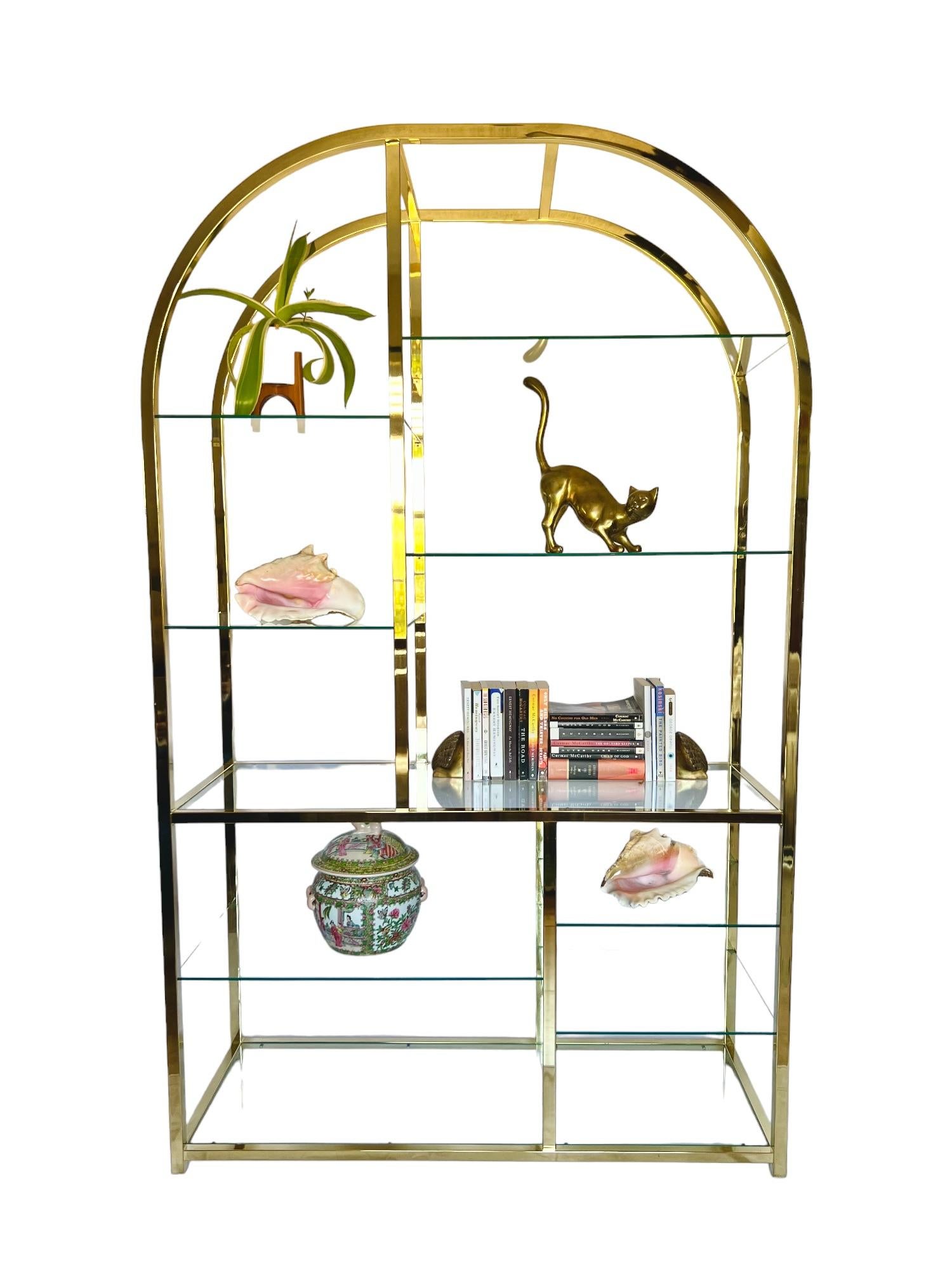 A vintage Hollywood regency style polished brass plated metal étagère featuring an arched design and nine glass tiered display shelves - two full-length, three medium & four small.
By DIA - Design Institute of America, 1985.

Dimensions: 48
