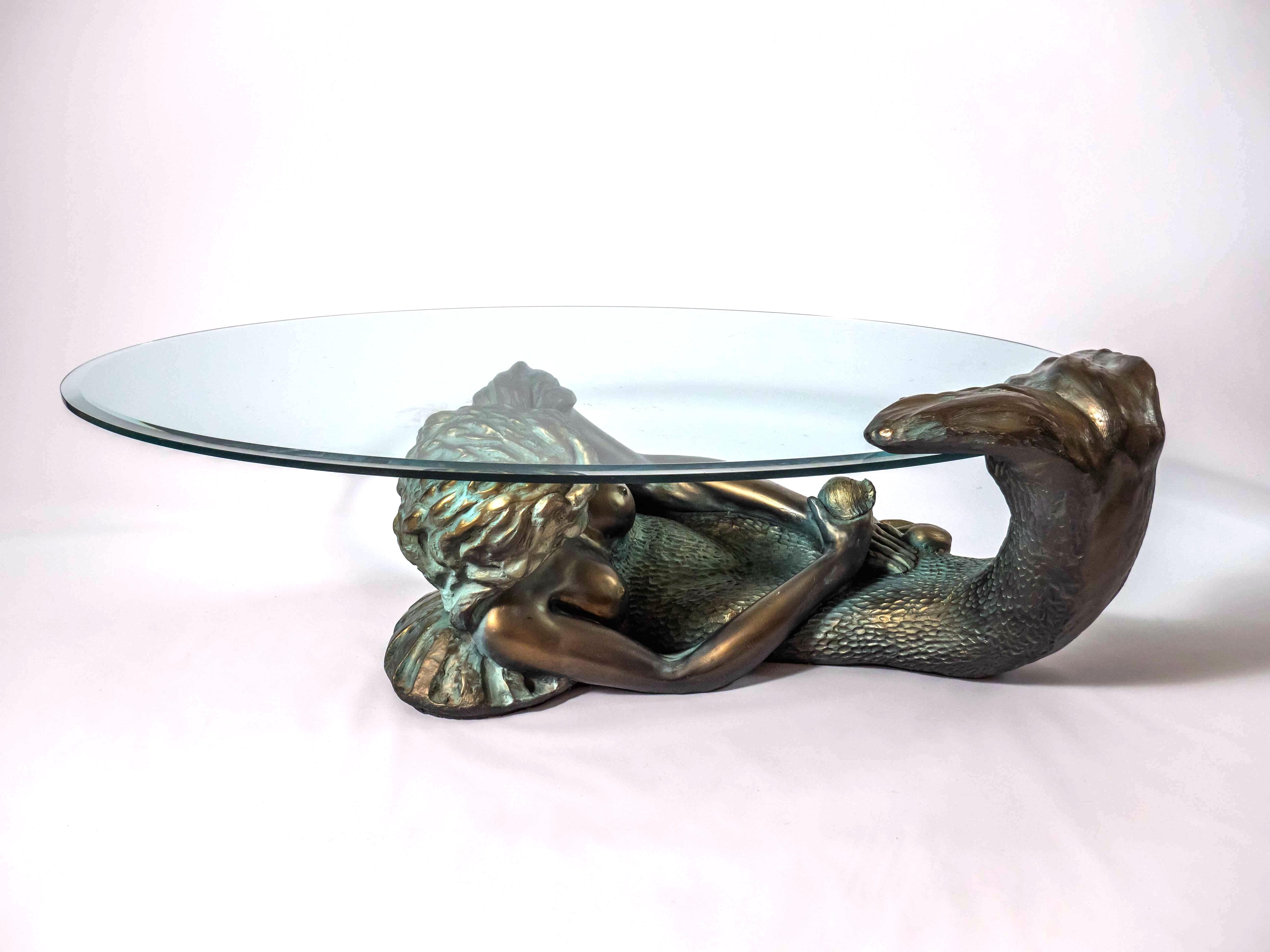 A beautiful coffee table made of a statue of a mermaid carrying a glass top. Heavy and stable table. Statue of a beautiful young mermaid. The top plate is oval and has a faceted edge. There is a signature on the back. The statue is made of epoxy