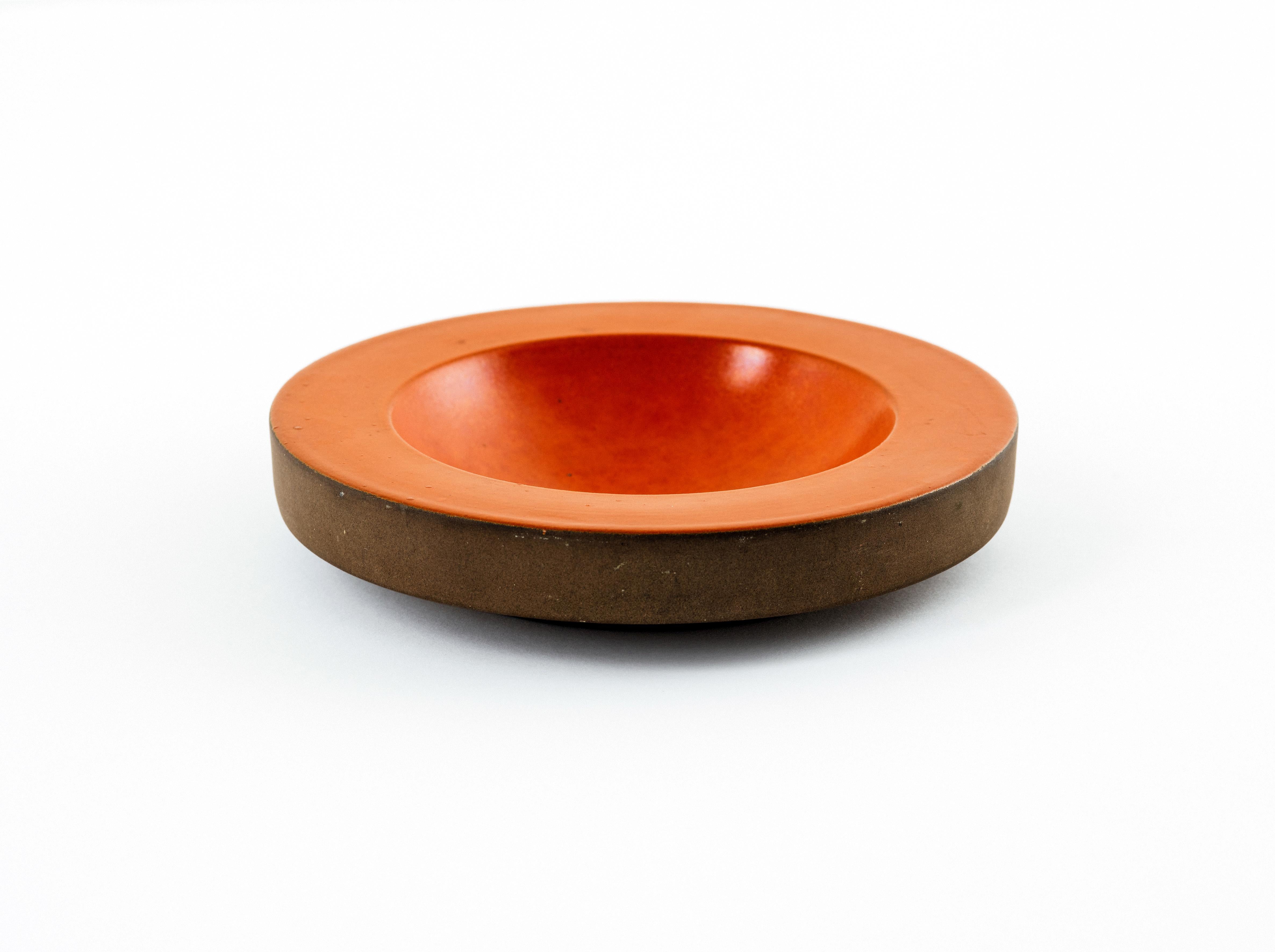 Modernist vide poche (catchall) or low bowl in slip-cast ceramic with a satin, blood orange glaze to the interior and rim, its edge and underside left unfinished for a striking contrast in color, sheen, and texture. The body floats over a recessed