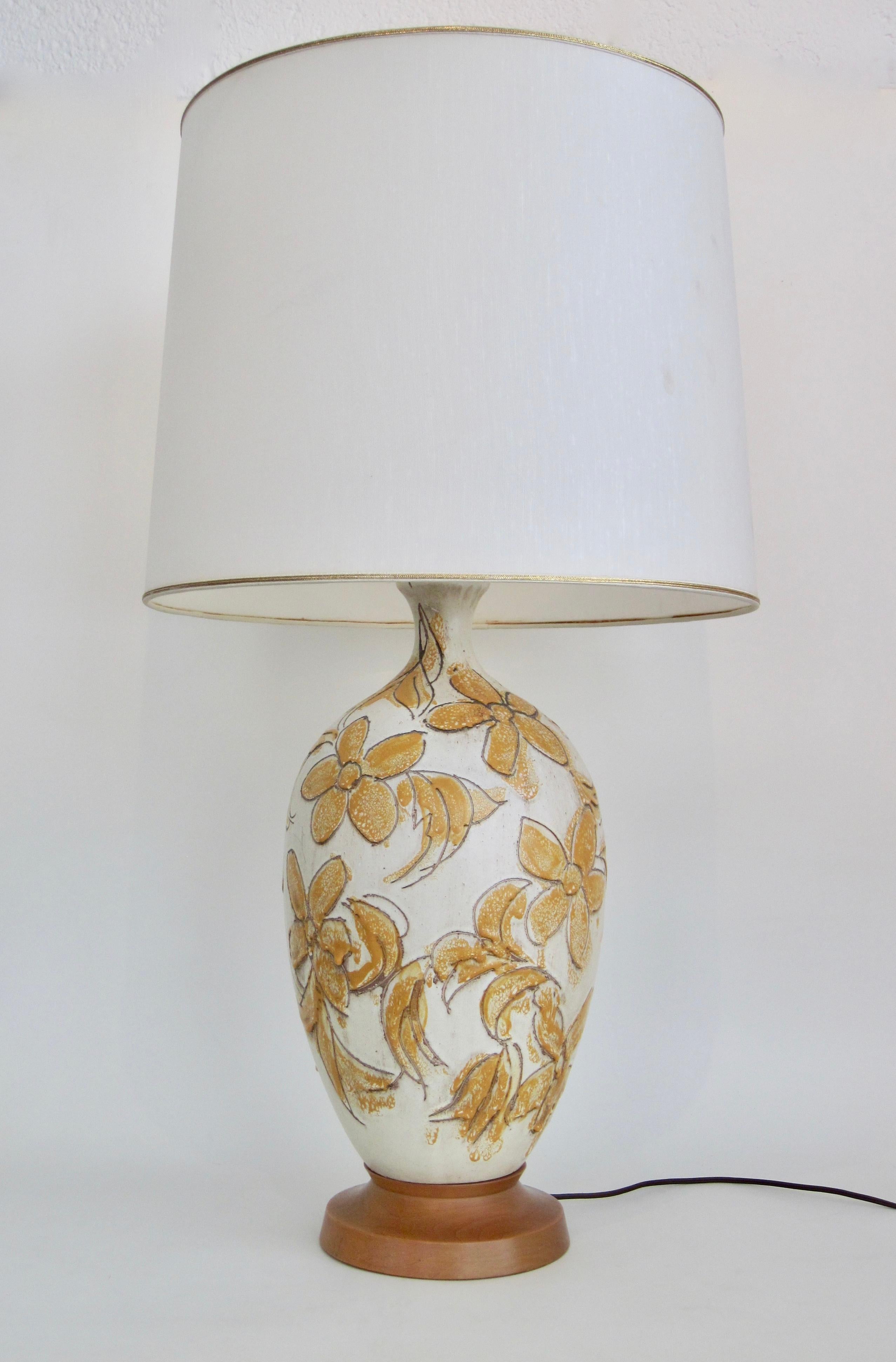 Cette lampe de table en grès présente des fleurs incisées sur une forme de goulot en grès blanc cassé de 21 pouces sur une base en bois. La couleur jaune moutarde peinte au pinceau et la glaçure goutte à goutte en font un motif distinctif pour