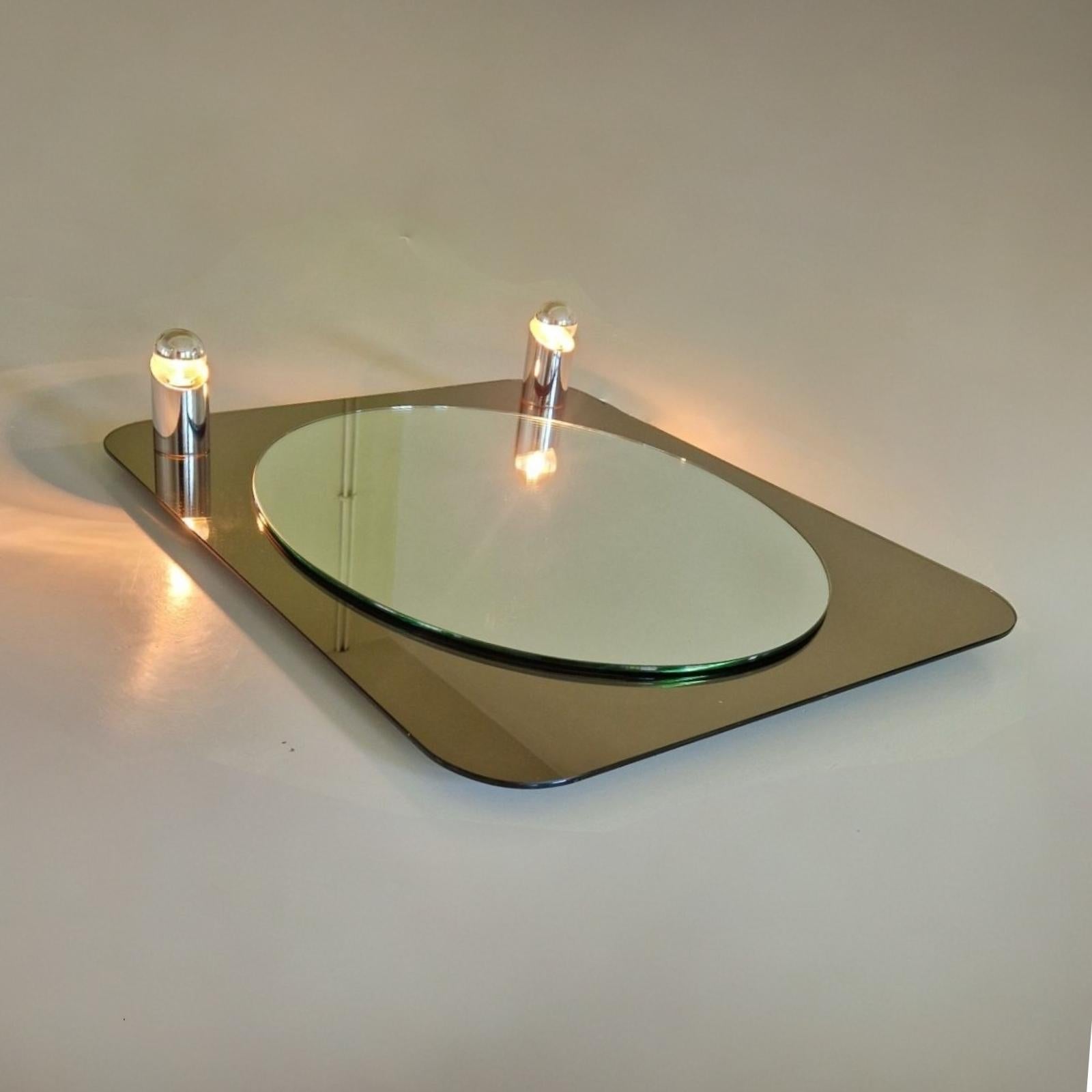 Bei diesem Designspiegel ist ein ovaler Spiegel auf einen rechteckigen Spiegel montiert. Die Rückwand ist mit zwei verchromten Lampen ausgestattet, die im Glas befestigt sind.
Die Kombination aus klarem und bronziertem Glas mit dem Chrom der Lampen