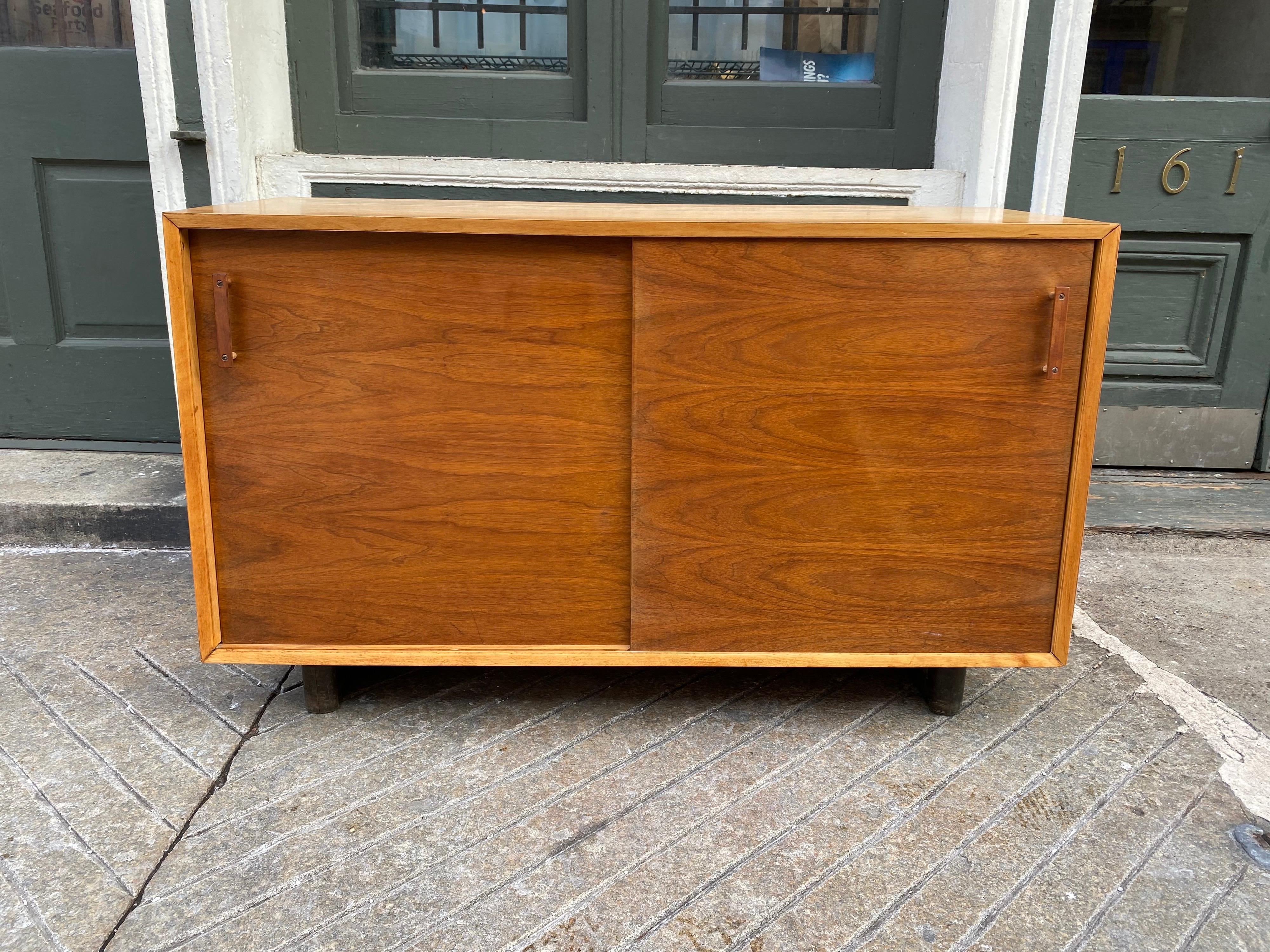 Designcraft sliding 2-door cabinet with a birch outer cabinet and walnut doors. Doors slide open to 1 shelf on each side. Original condition shows normal wear, slight shadow to top where something sat. Two plinth legs support the cabinet. Unusual