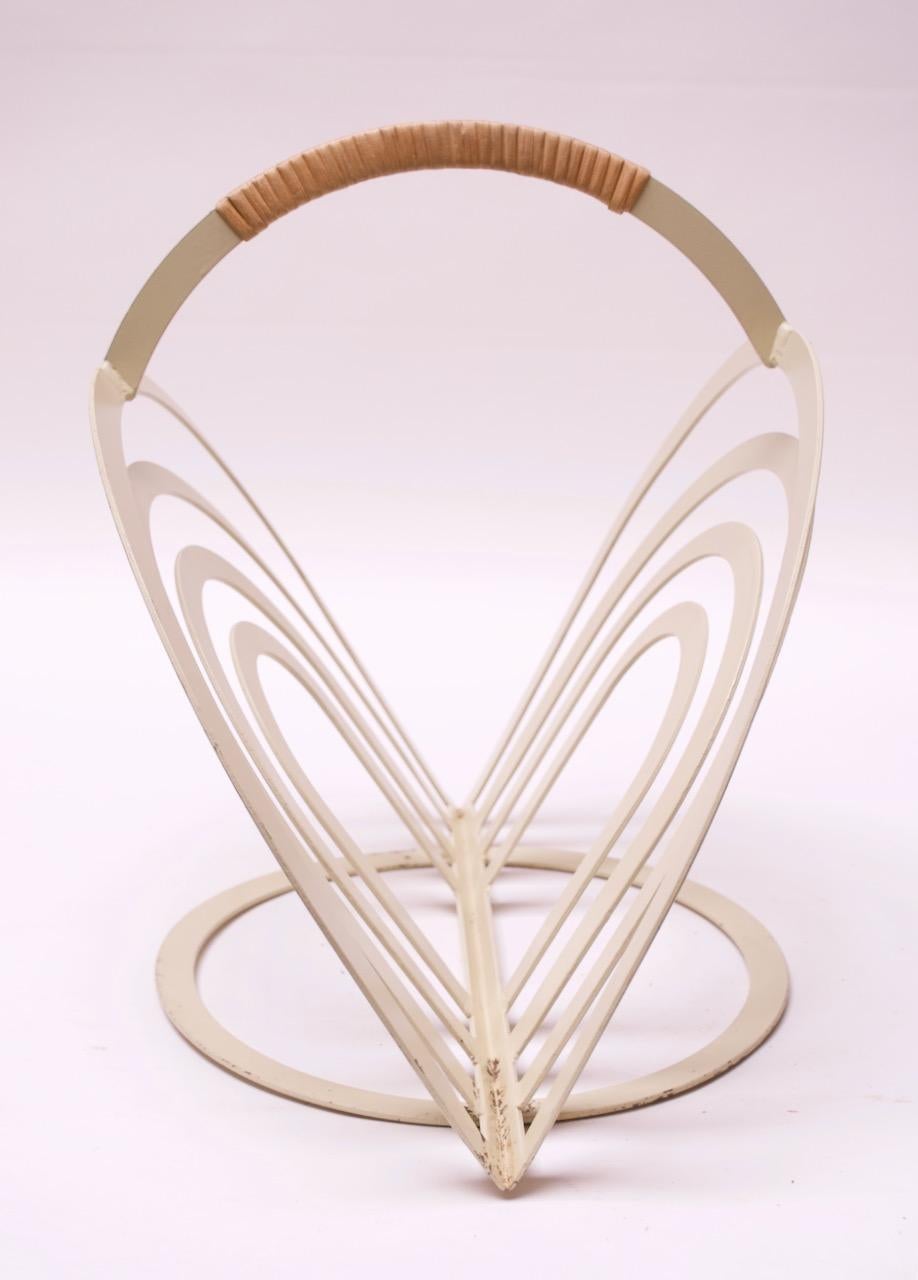 Magazine rack in off-white painted metal with a cane-wrapped handle, circa 1970s. Features two splayed 'arches' supported by a round base. Very unique form, reminiscent of designs by Vernor Panton, but artist is unknown.
Paint loss as shown to rack