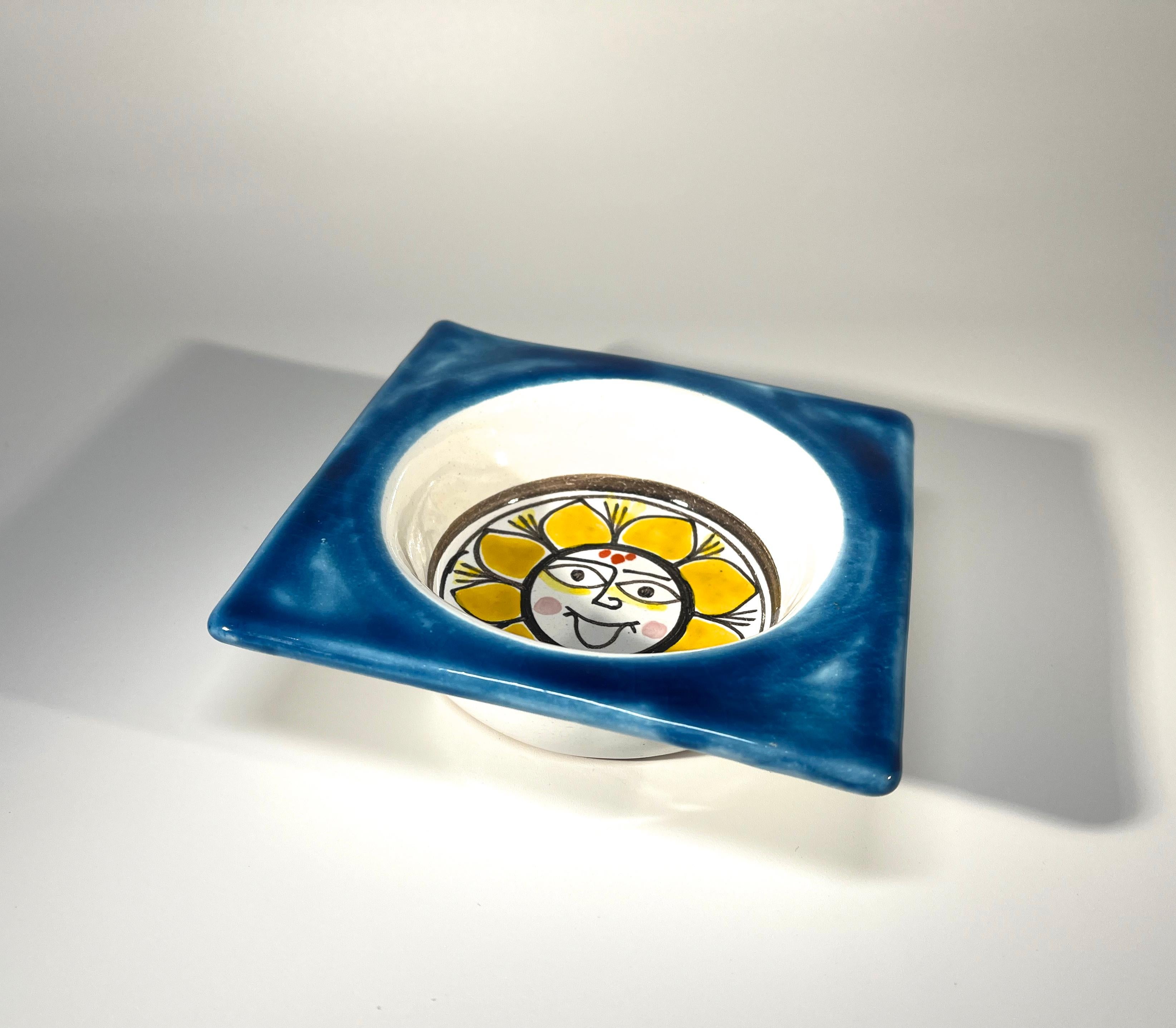 DeSimone of Italy, 'Sunny Sunflower'  is typical of DeSimone's colourful and playful art.
Hand painted yellow sunflower motif to centre with a bright blue rim
This small bowl is weighty and almost square -  a super little piece for the DeSimone