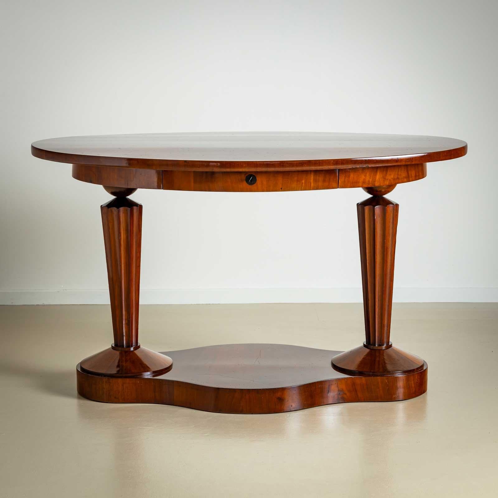 Biedermeier Schreibtisch mit ovaler Tischplatte und geschwungenem Sockel. Die Beine bestehen aus konischen, kannelierten Säulen mit Kugelkapitellen. In den Rahmen ist eine Schublade integriert. Der Schreibtisch ist in Kirsche furniert und wurde