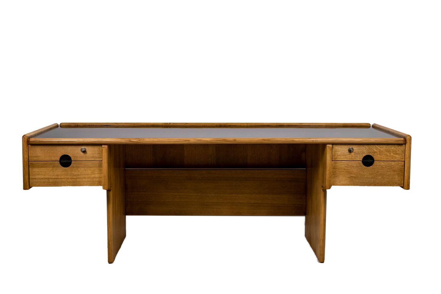 Large desk in oak, rectangular in shape. Black tray standing on two boxes with two drawers, opening with circular and semi-circular shaped handles. Keys.

French work realized in the 1970s.