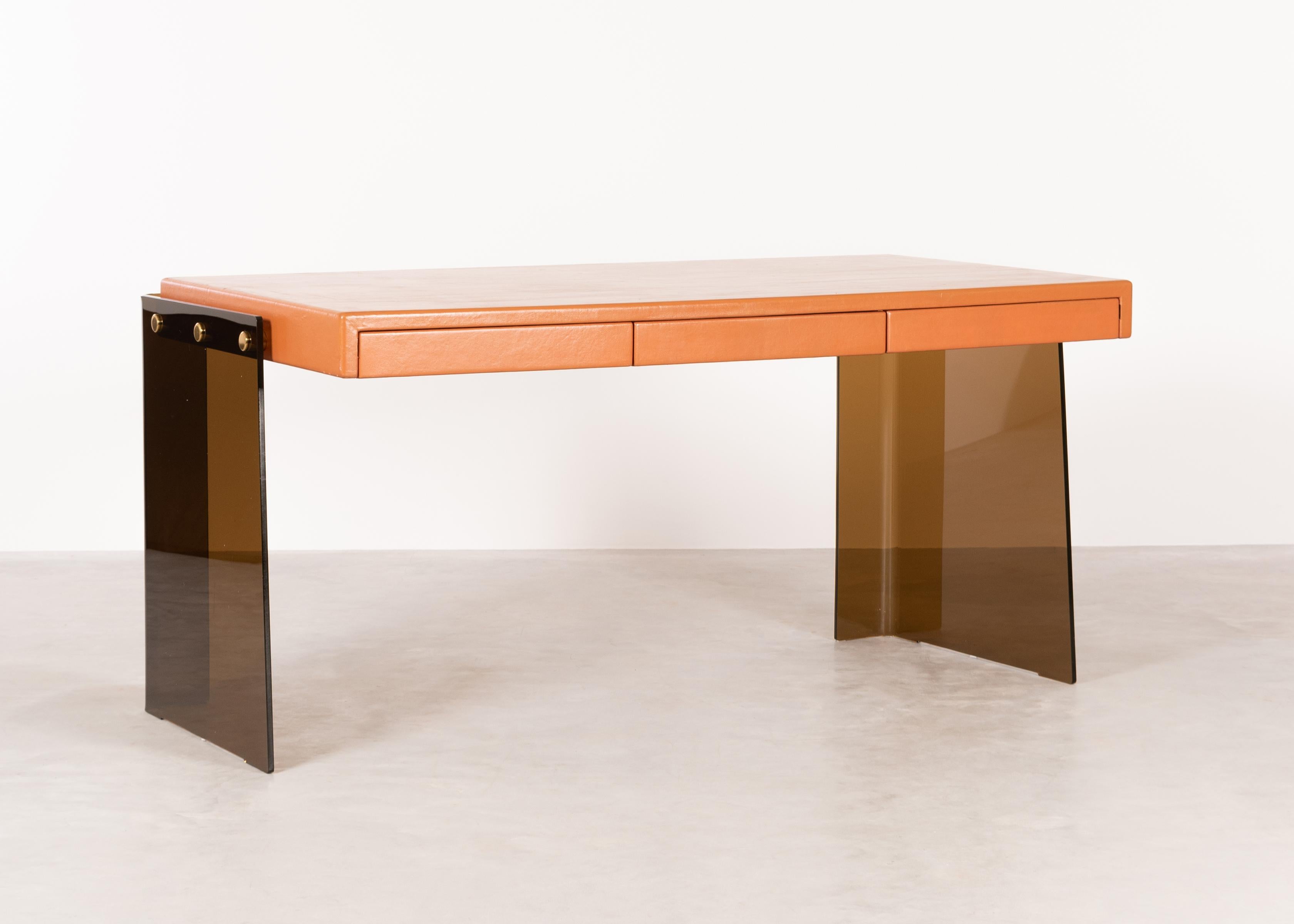 Stylish desk by an unknown designer (in style of Michel Dumas) made in the mid-1970s in France. Frame of smoked plexiglass and tabletop (joined with brass pins) upholstered in orange leather containing 3 drawers. All in very good condition with