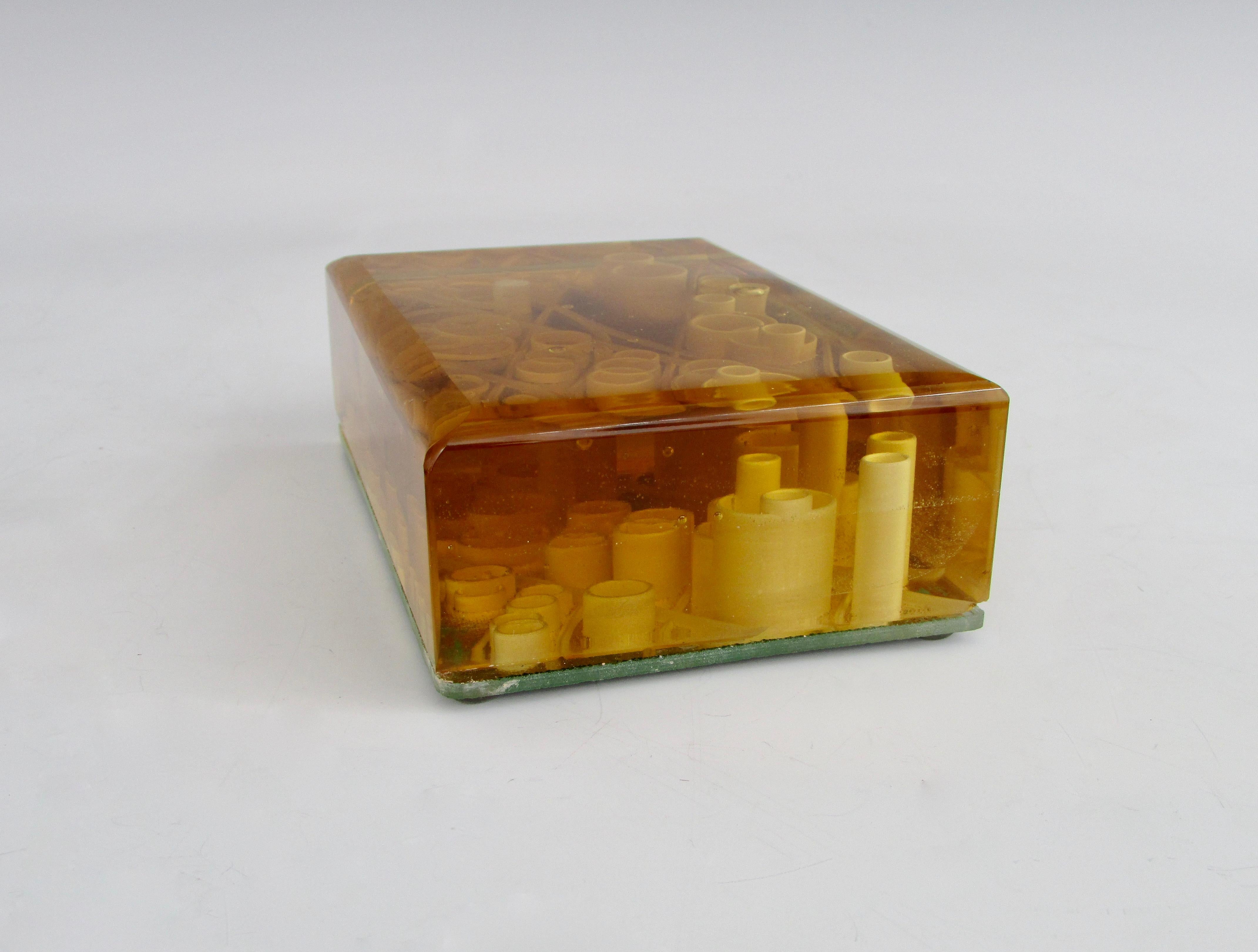 Various size cylinder pieces set in amber tone glass. Sculpture is set on top of a mirrored base. Illegible signature on one side.