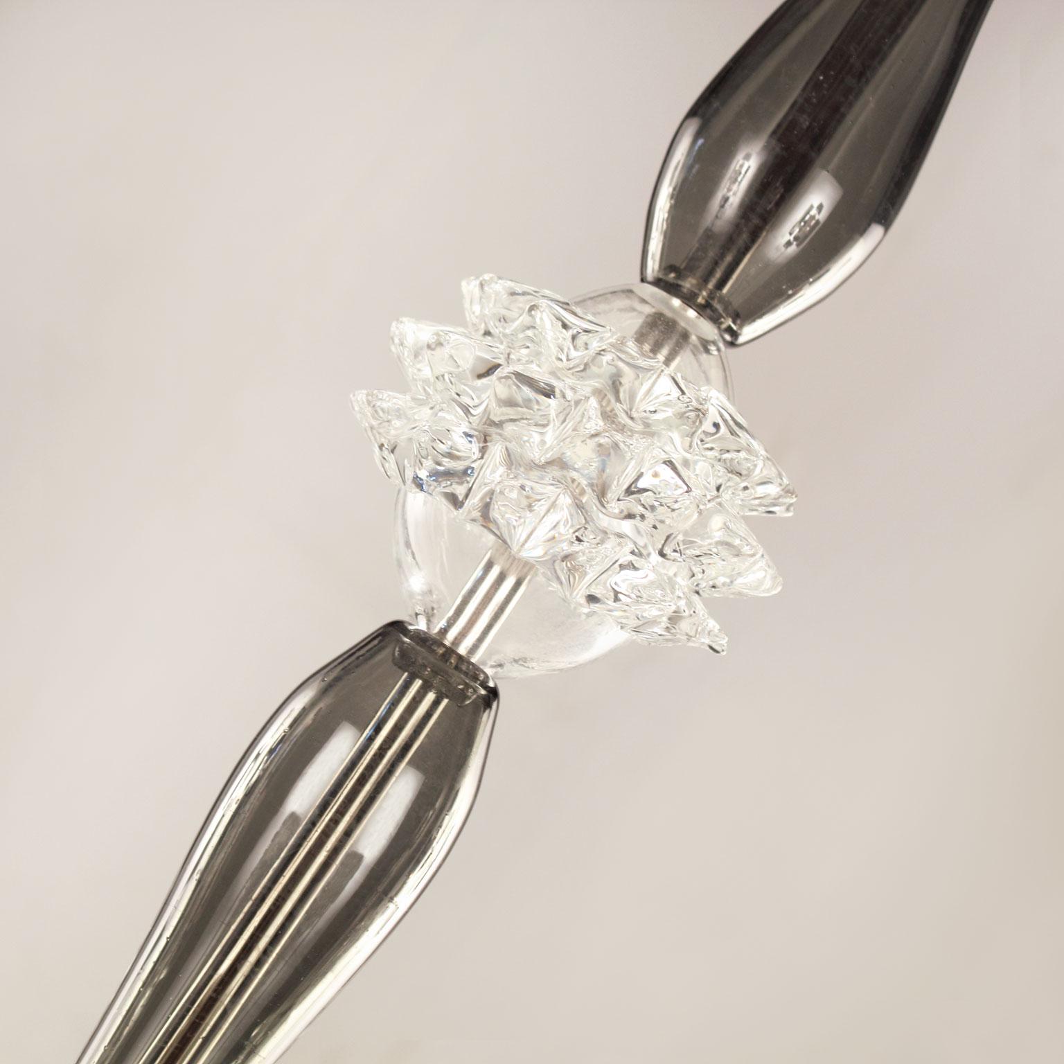 Die mundgeblasenen Glaskronleuchter Diamante von Multiforme zeichnen sich durch ein schlankes zentrales Element aus.
Die Arme, die Mittelsäule und der Abschlussbecher sind aus makellosem, glattem Glas gefertigt. Die herausragenden Elemente dieses