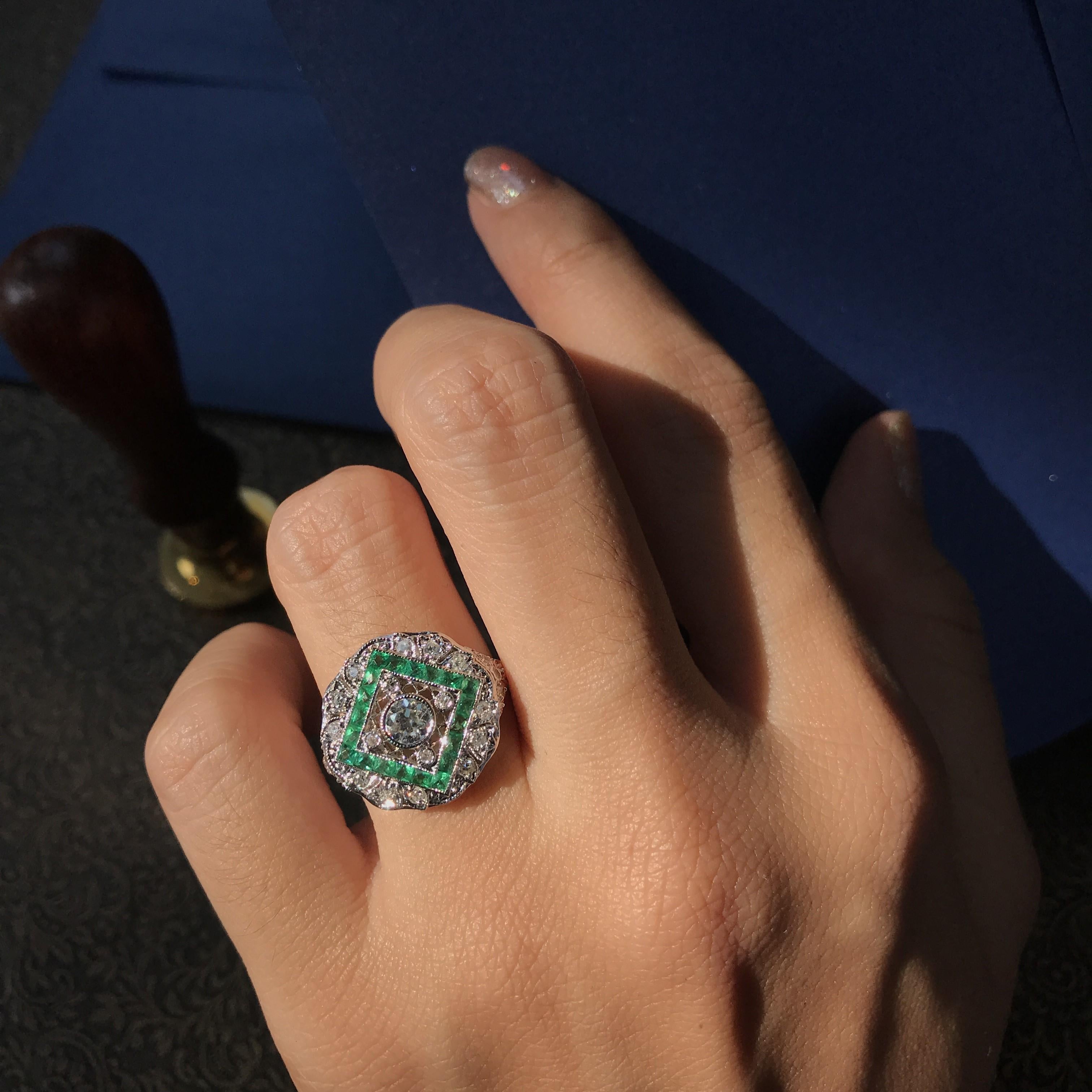 An Art Deco-style engagement ring showcases a brilliant diamond at its center, radiating timeless elegance. Surrounding the centerpiece is a striking square-line arrangement of emeralds, adding a vivid pop of color and vintage charm. The final outer