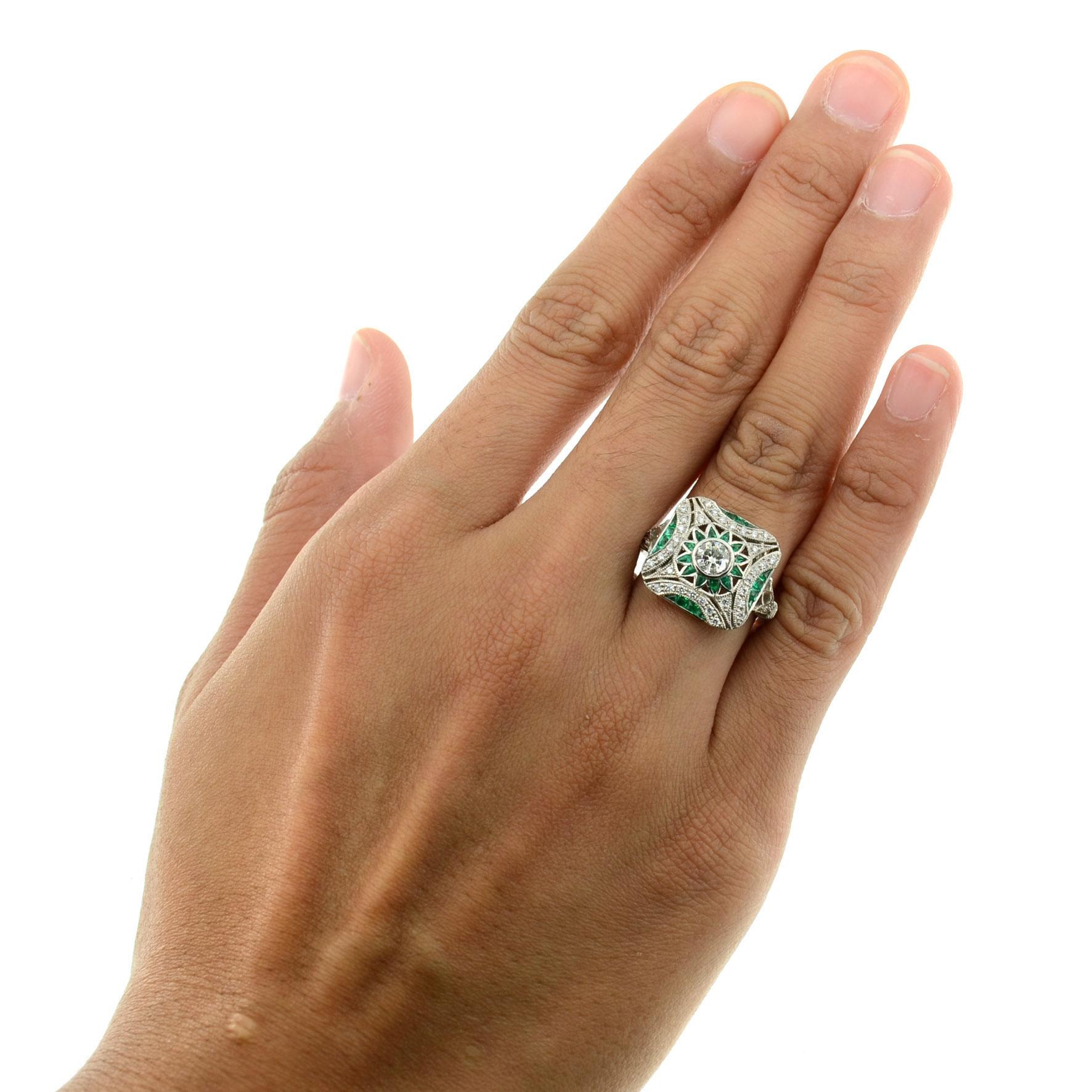 This vintage style ring centers a round cut diamond, weighing approximately 0.50 carat in bezel set. The center stone is accented by a floral halo of vivid green emerald and round white diamonds. A border of diamonds is set along the edge of the