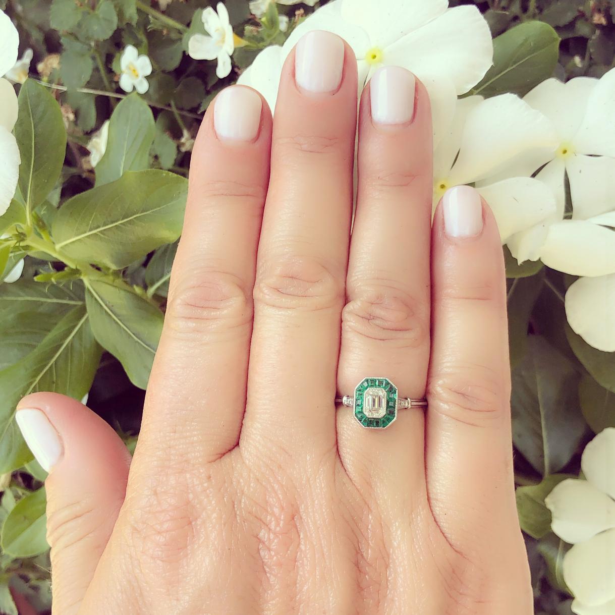 Go back to an era of fine design with this lovely Art Deco inspired 18k white gold ring! It centers a bezel-set emerald-cut diamond, weighing an estimated 0.50 carat, G/VS, set within a frame of calibre-cut emeralds, and then highlighted with 4