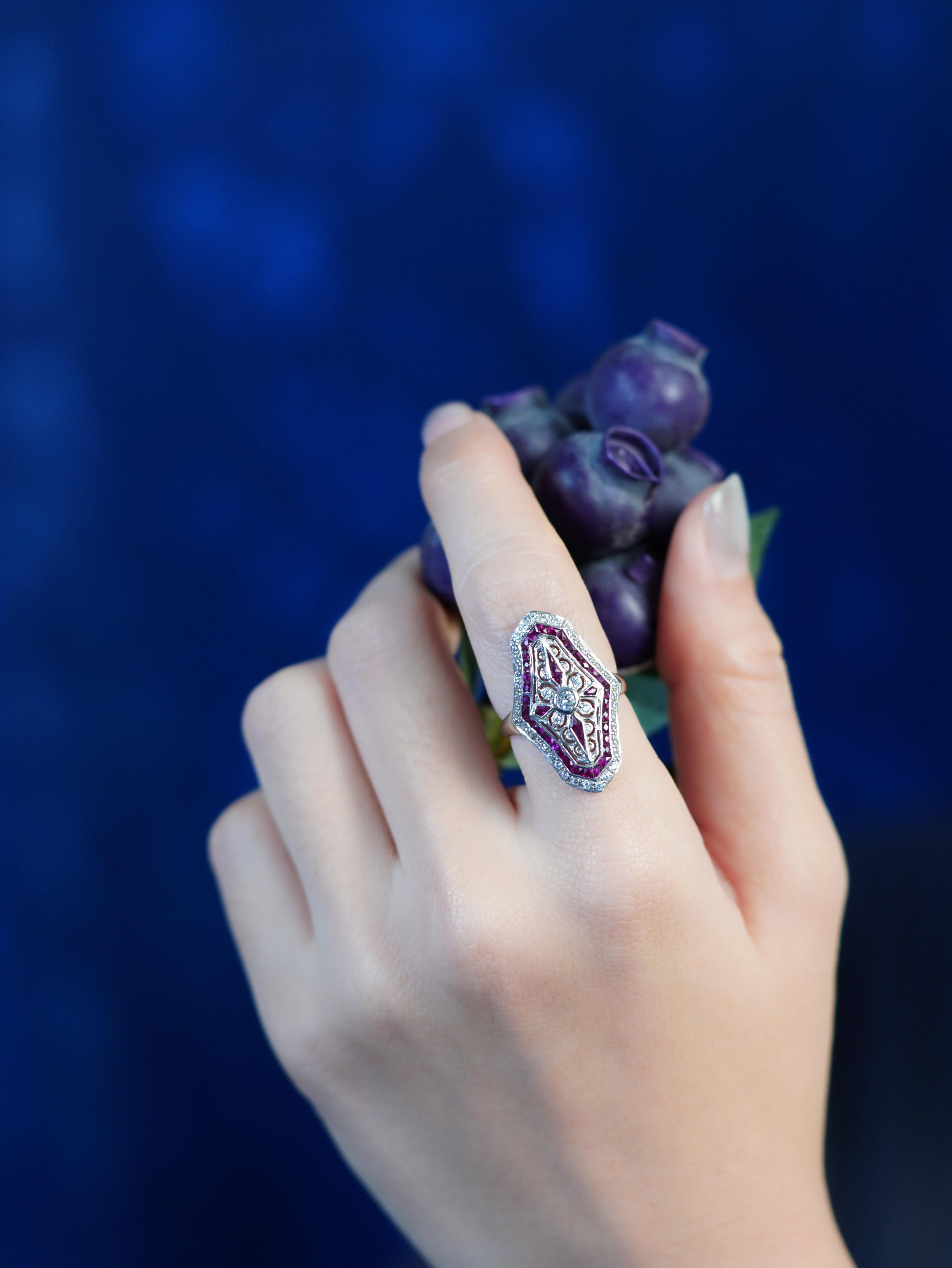 Un diamant étincelant occupe le devant de la scène, inspiré par de délicats motifs floraux. Des rubis taillés en France encerclent la pierre précieuse, ajoutant une riche couleur et une élégance géométrique. La couche extérieure de diamants rehausse