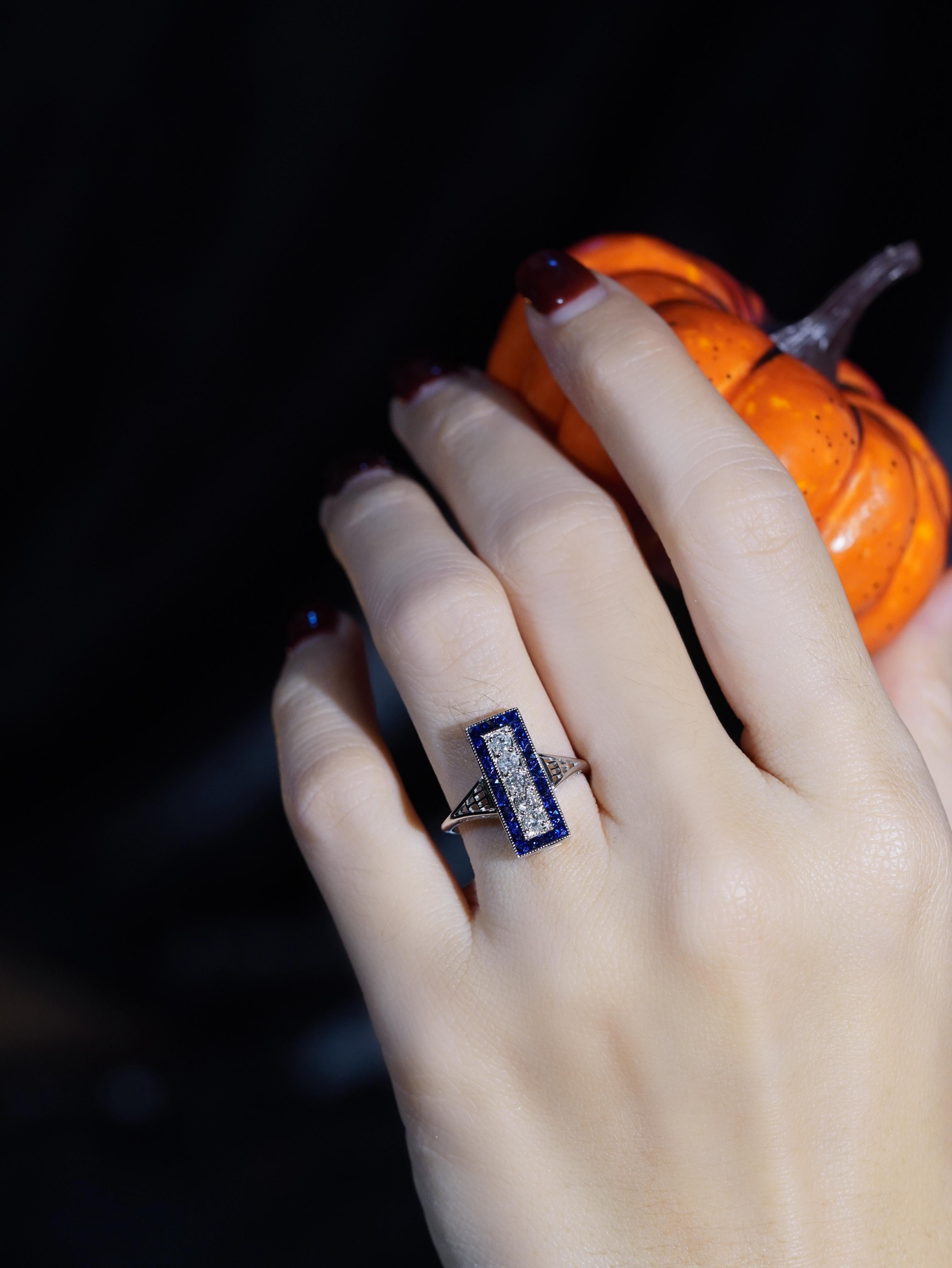 Un vero simbolo di eleganza senza tempo. Cinque diamanti rotondi brillanti siedono con orgoglio al centro, assicurati da griffe e incorniciati da un'audace aureola di zaffiri taglio francese che aggiungono un contrasto ricco e regale. Le spalle con