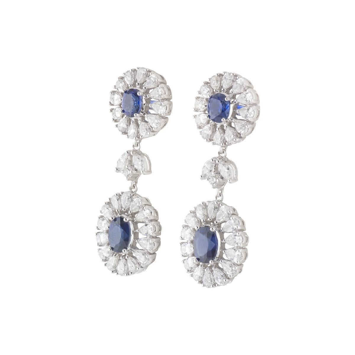 A sparkly pair of 18k white gold diamond and sapphire drop earrings. The earrings each comprise of an open work circular motif with an oval cut sapphire and a halo of 12 pear cut diamonds. Complementing the motif is another open work oval motif with