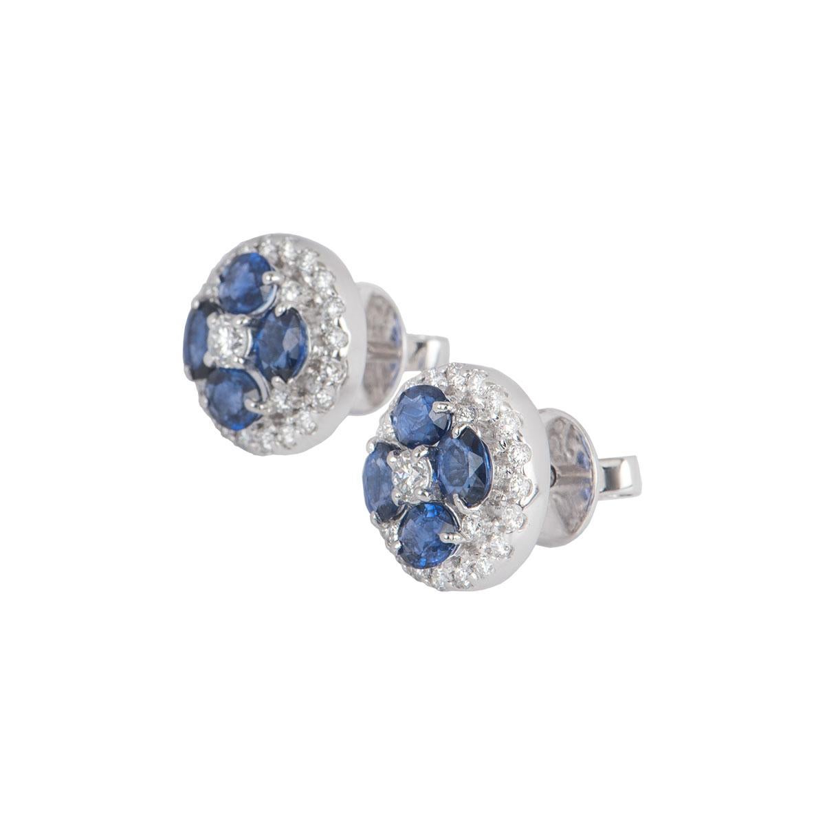 A pair of 18k white gold diamond and sapphire earrings. Featuring a floral design with a round brilliant cut diamond in the centre surrounded by four oval cut sapphires with a halo of round brilliant cut diamonds. The diamonds have a total weight of