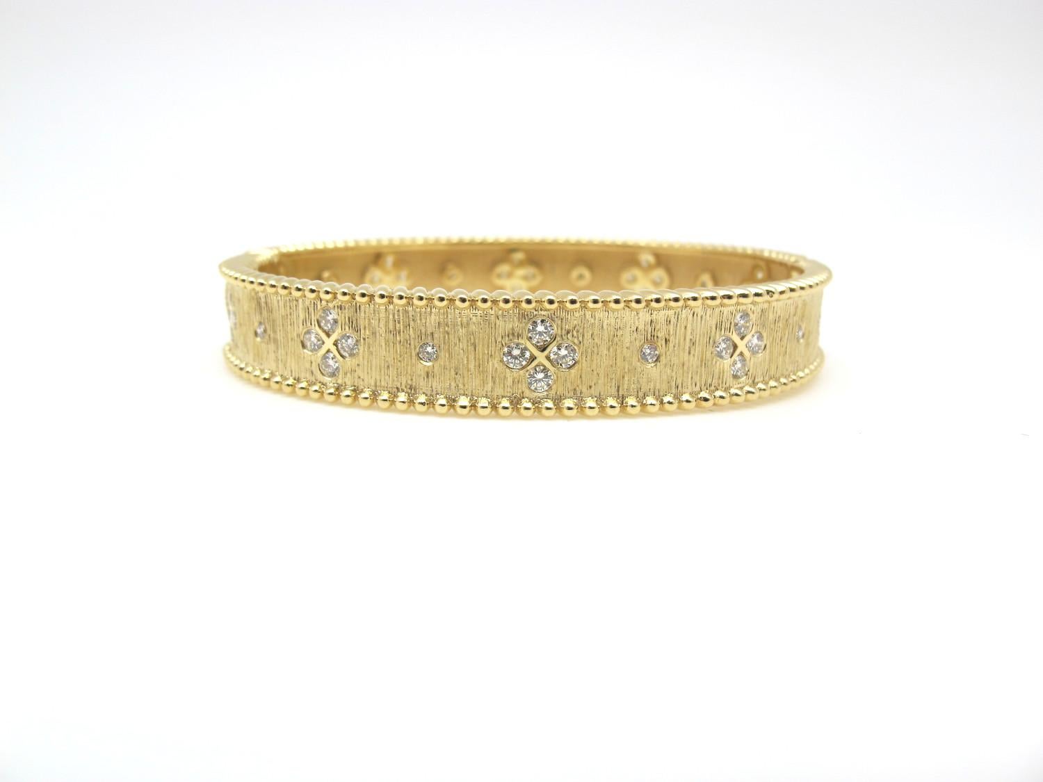 This elegant 18k yellow gold hinged bangle bracelet is beautifully textured and features 48 round brilliant cut diamonds that have been flush set in delicate floral motifs. Both sides of the bangle are adorned with a raised, decorative edge. The