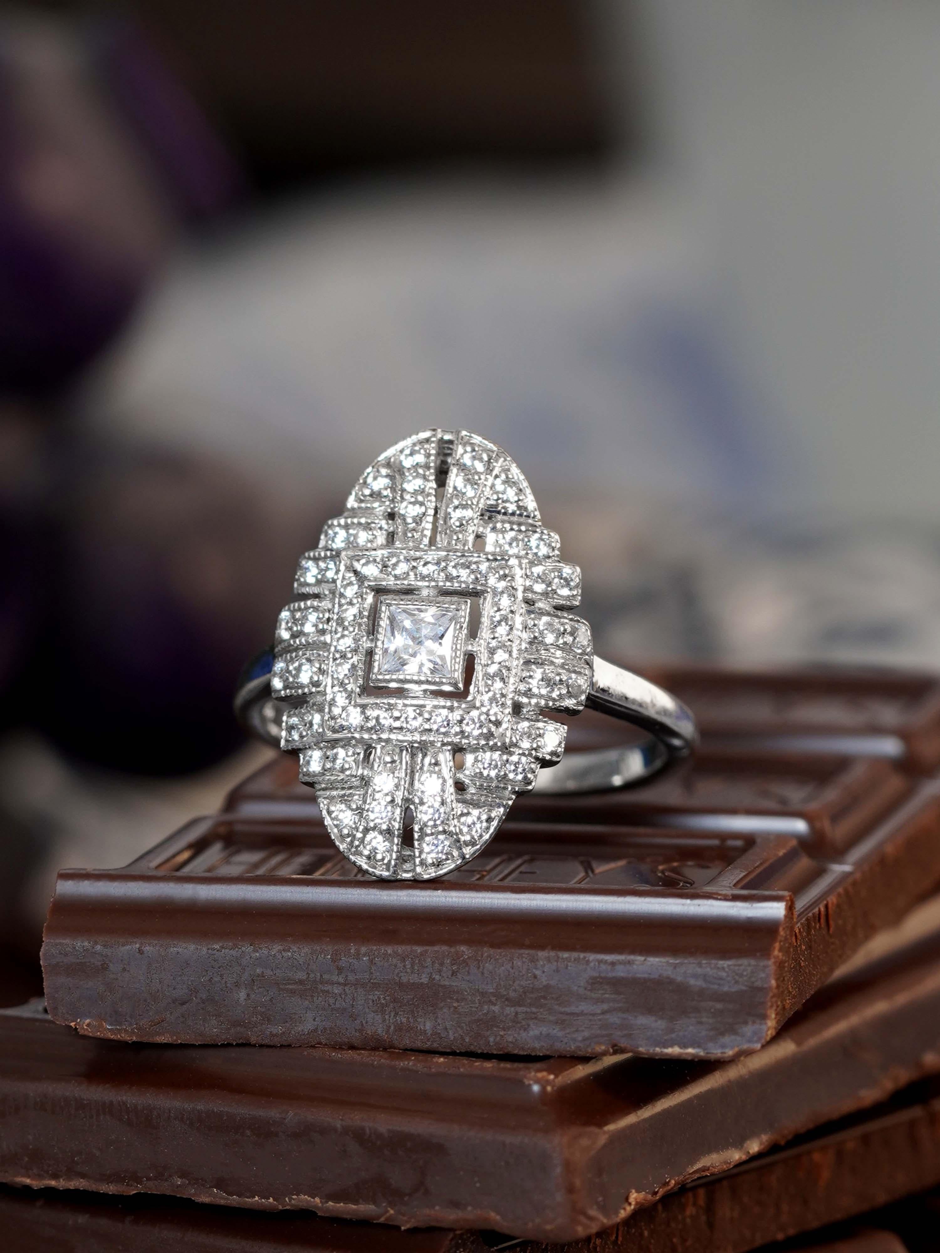 Un diamant taillé en carré domine le centre, scintillant avec une précision géométrique. Des lignes épurées et une symétrie audacieuse reflètent l'élégance intemporelle de l'Art déco. Les détails qui l'entourent rehaussent l'éclat du diamant, créant