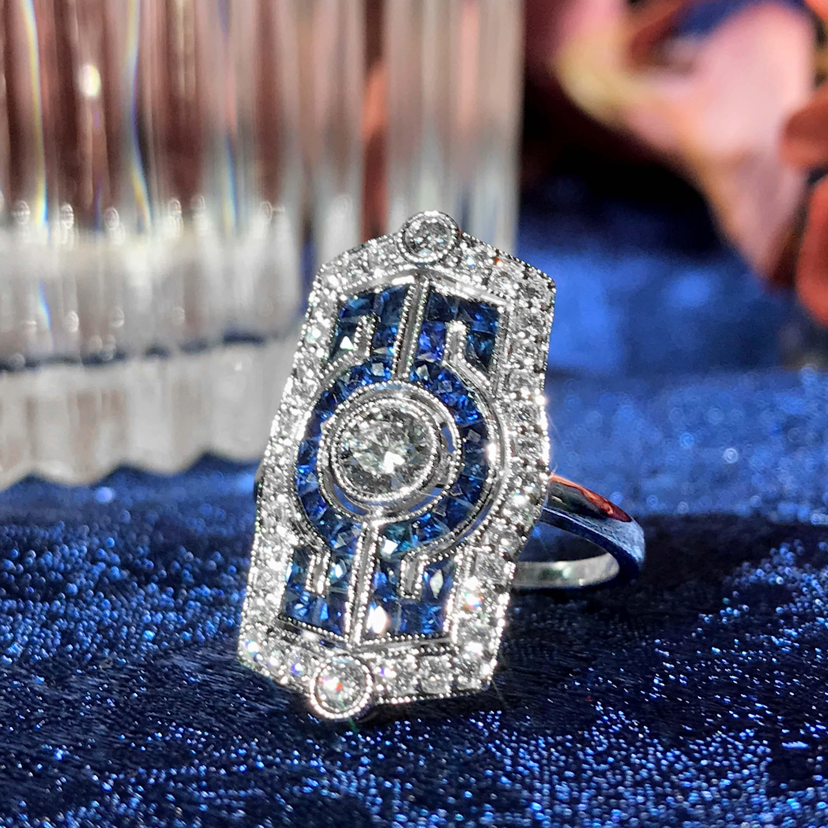 Cette bague d'inspiration Art déco accroche le regard grâce à ses diamants et ses saphirs bleus en or blanc étincelant. Un seul diamant rond de 0,30 carat taillé en brillant orne le centre de la bague. Les saphirs bleus de taille française