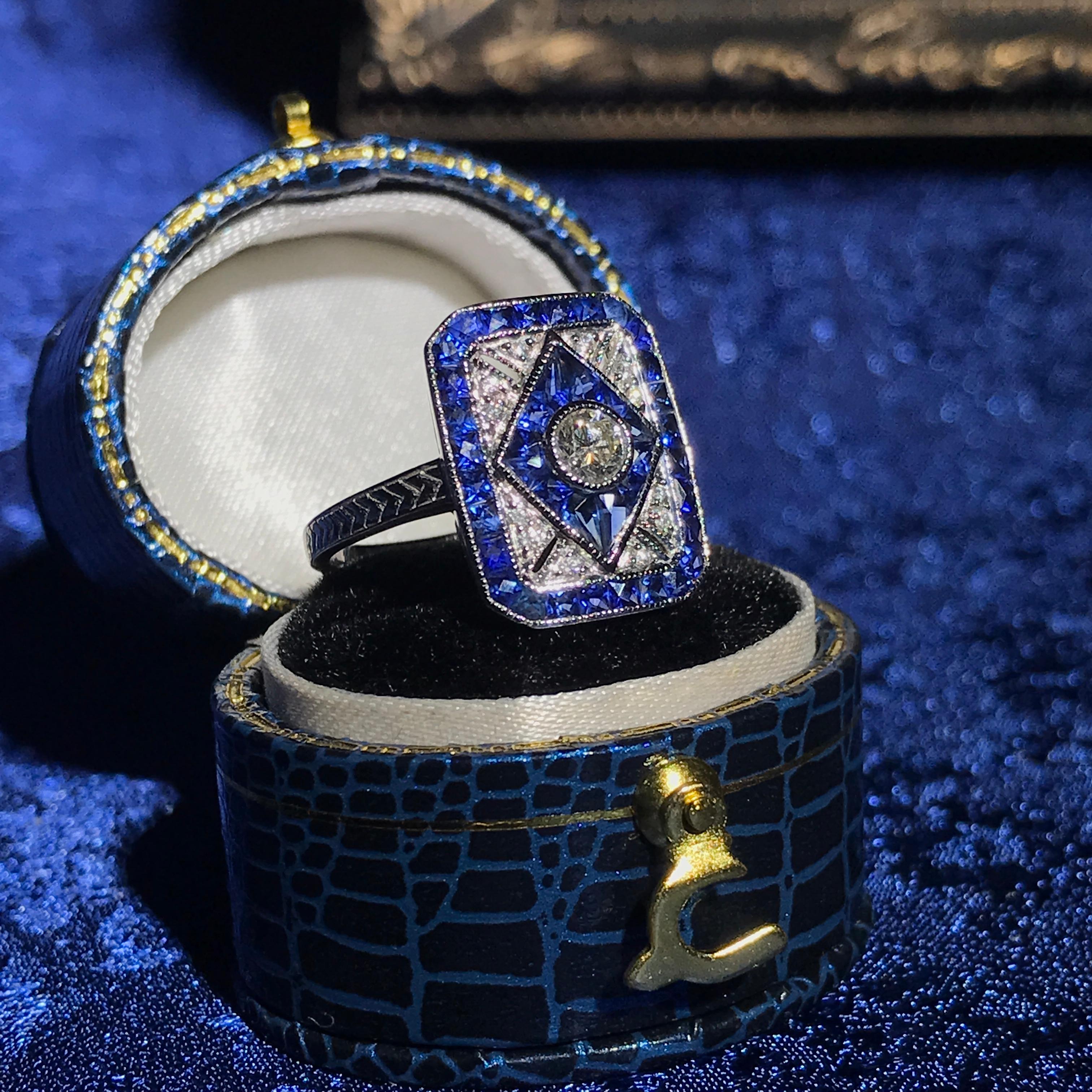 Celebrate your love story with this exquisite Diamond Blue Sapphire Art Deco Style Engagement Ring. At its heart lies a brilliant center diamond, elegantly showcased and framed by stunning French cut blue sapphires arranged in a unique rhombus