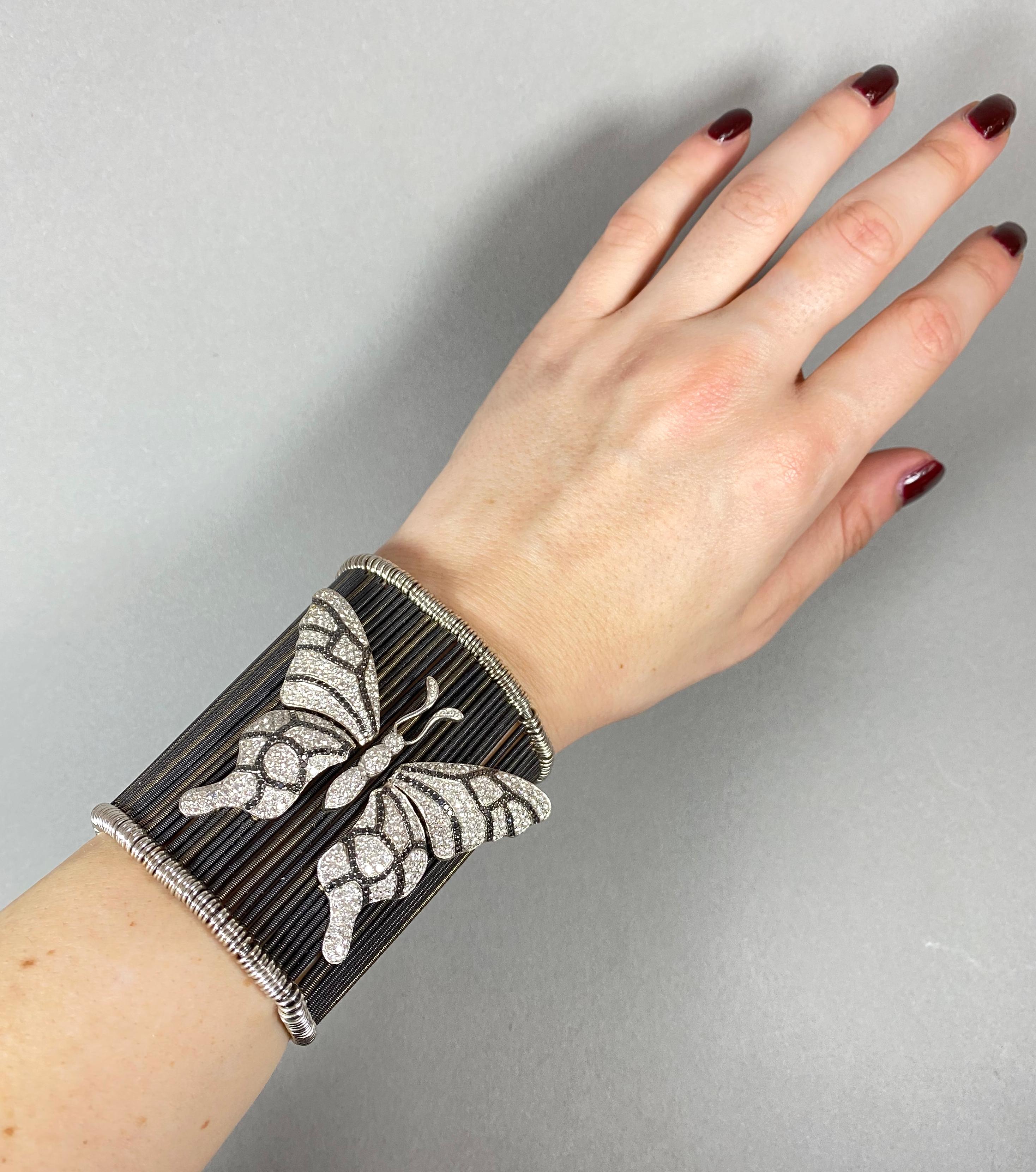 Ein modernes Manschettenarmband, bestehend aus eloxierten Weißgold-Drehstäben, auf gefederten Beschlägen, mit einem großen, mit weißen und schwarzen Brillanten besetzten Schmetterling auf der Vorderseite, mit zwei kleineren Marienkäfern auf der