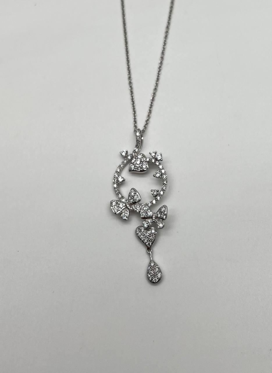 Découvrez la beauté raffinée de ce collier à pendentif. Cette pièce exquise se compose d'une chaîne délicate menant à un pendentif orné de diamants pavés étincelants. Le motif circulaire est agrémenté de cœurs et de nœuds et se termine par un