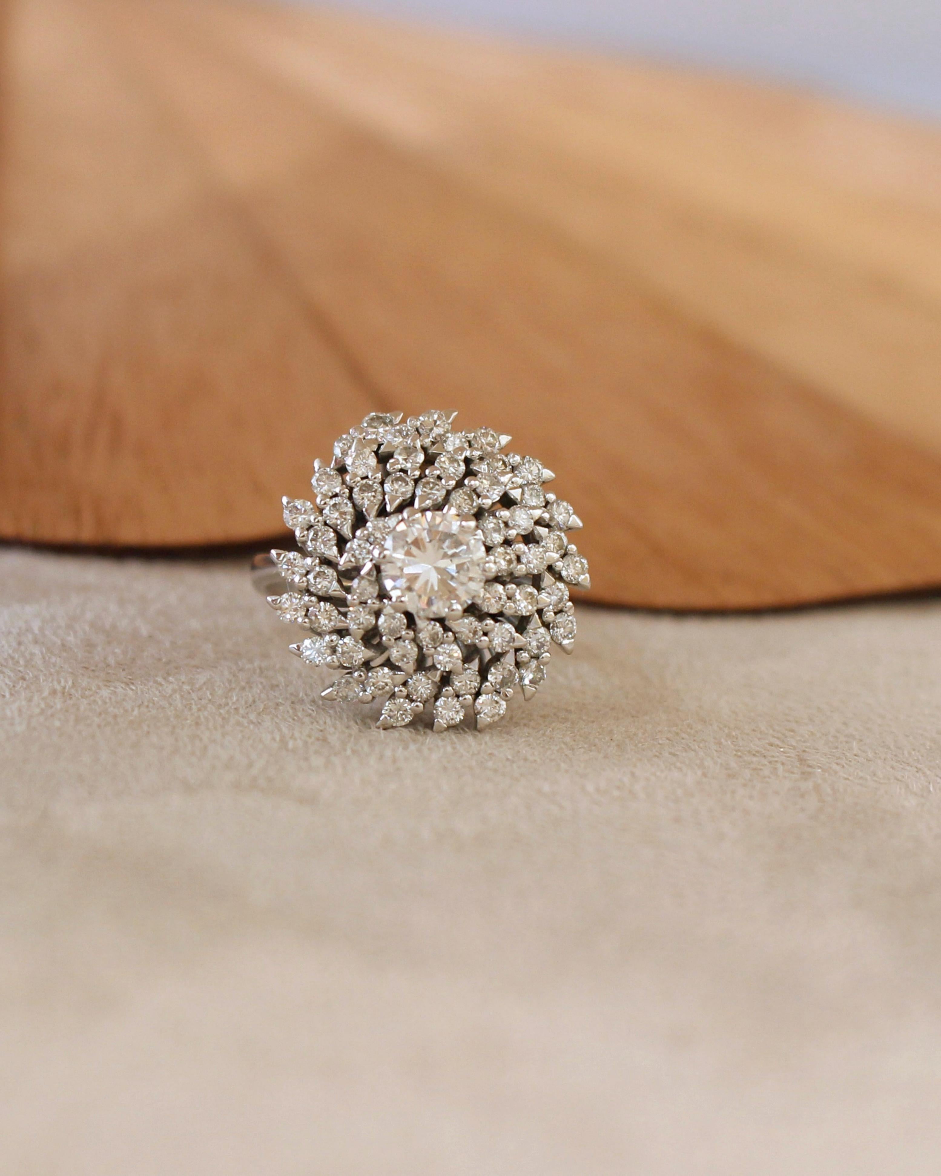 Anillo de cóctel de oro blanco de 18 quilates decorado con un diamante central talla brillante de 0,90 quilates engastado con 6 garras. La piedra central está rodeada por 58 diamantes redondos de 0,02 quilates cada uno. 
La cesta del anillo está