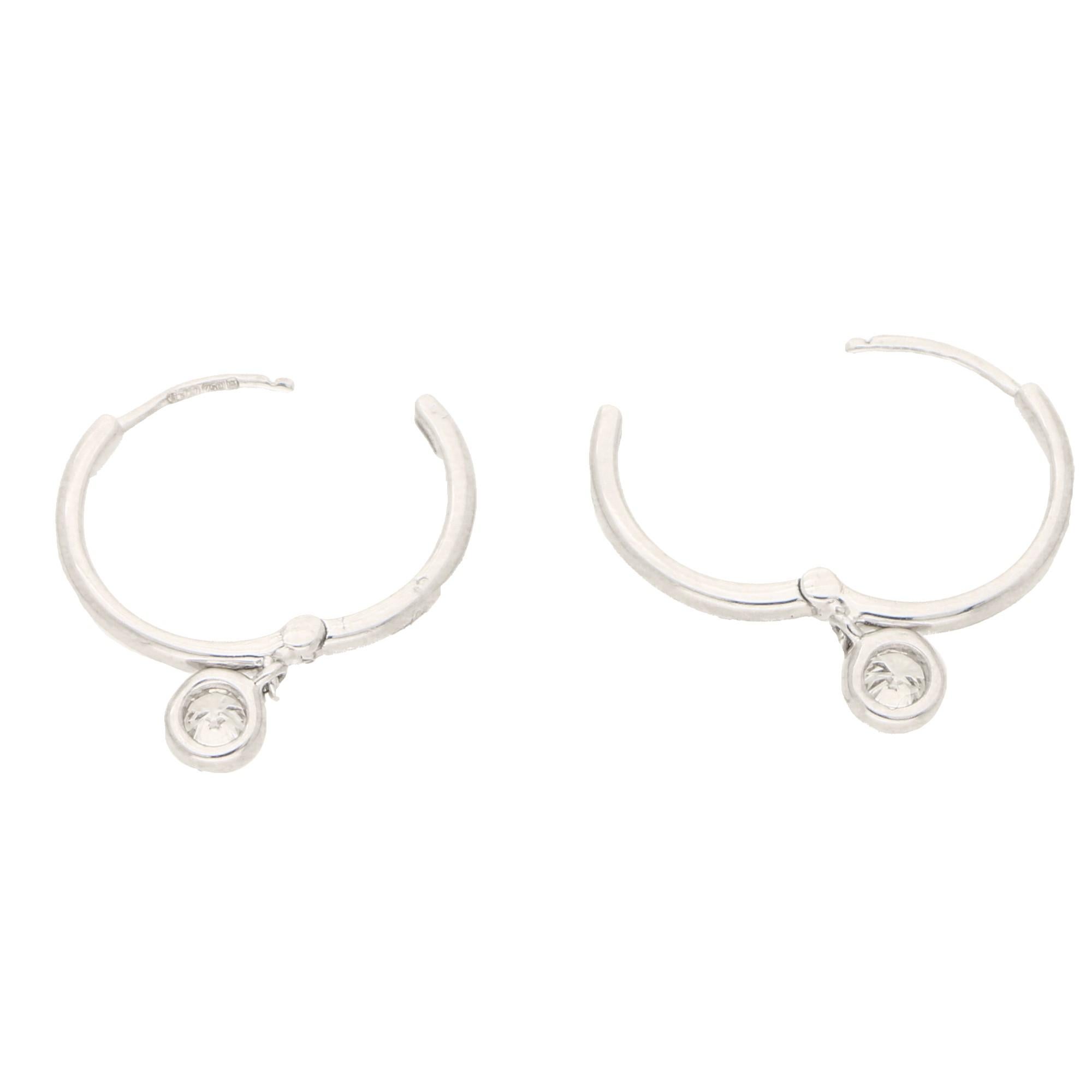 A lovely pair of diamond drop hoop earrings set in 18k white gold.

Each diamond is composed of a simple solid 18k white gold hoop. Suspended from the bottom of the hoop is an articulated rub-over set diamond drop which hangs elegantly from the ear.