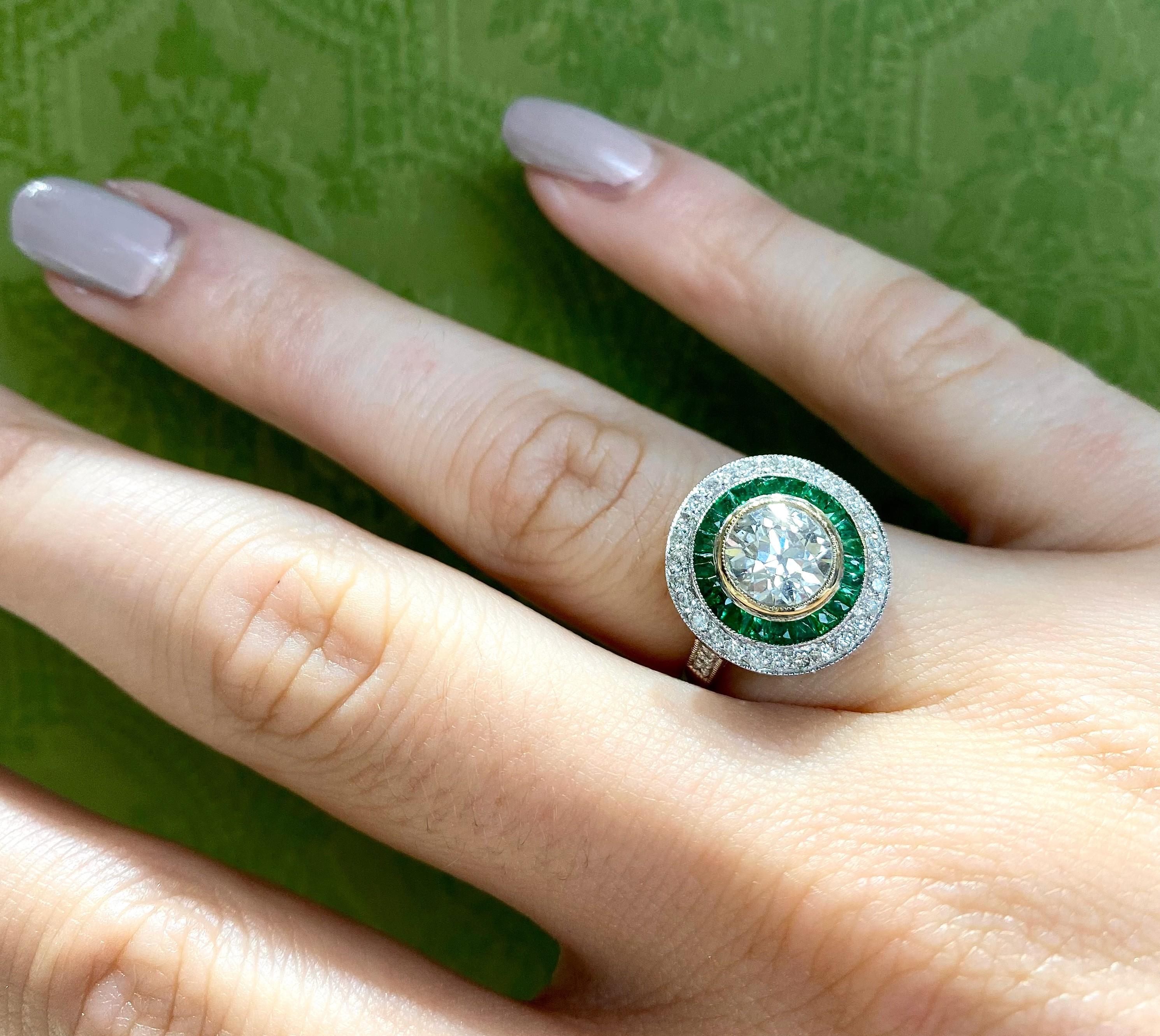 An Art Deco style diamond and emerald ring, set with a 1.29ct round European old-cut diamond, in our opinion the colour is K, our opinion of the clarity is I1, in a millegrain edged rub over setting, surrounded by twenty-two, French-cut emeralds, in