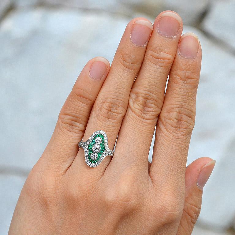 Glimmering with diamonds and drama, this vintage-inspired Art Deco style ring will have heads turning. Crafted in platinum, this shimmering look features three round diamonds wrapped in sapphire French cut emerald and diamond-lined halo frames