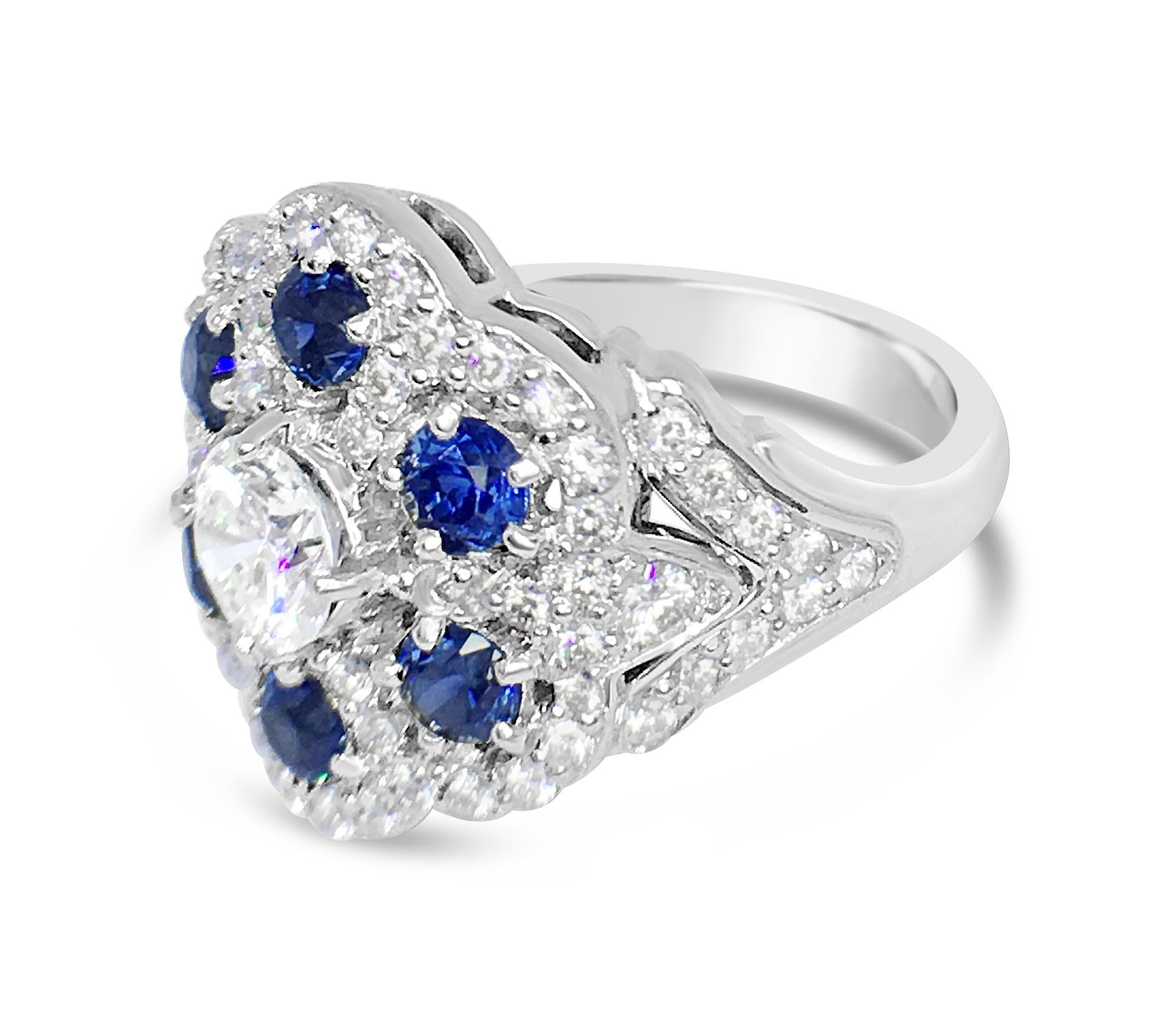 A beautiful and delicate 18 karat white gold ring set with a center diamond of 0.61 carats H color and VS clarity surrounded by 1.22 carats total of blue sapphires and another 0.69 carats total weight of diamond pave.  The ring is a size 6.5 and is
