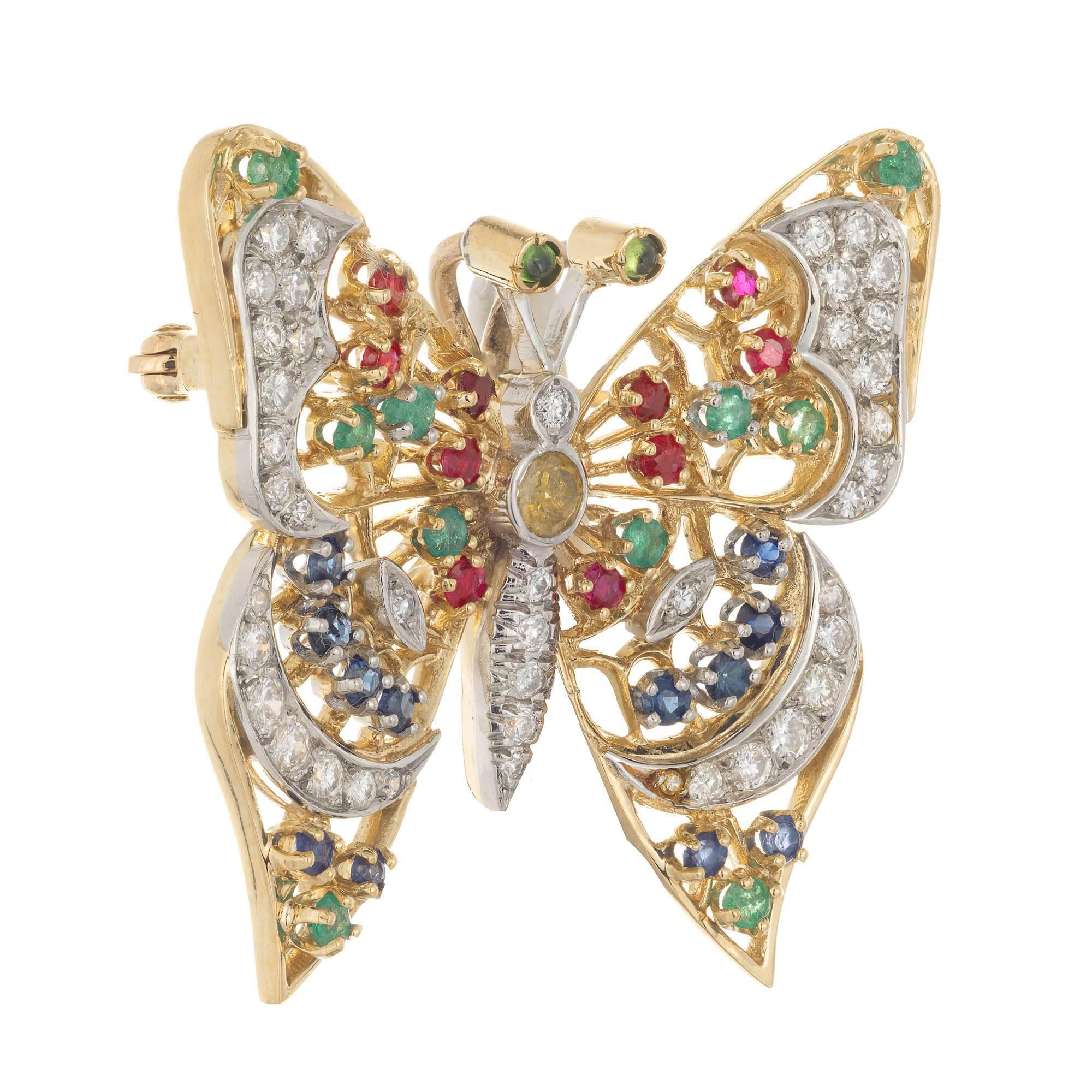 Yellow gold diamond and colored stone set butterfly brooch. Decorative butterfly has white pave set diamonds in each wing and down the center part of body.  Emeralds, rubies and sapphires are prong set in each wing with peridot cabochon bezel set in