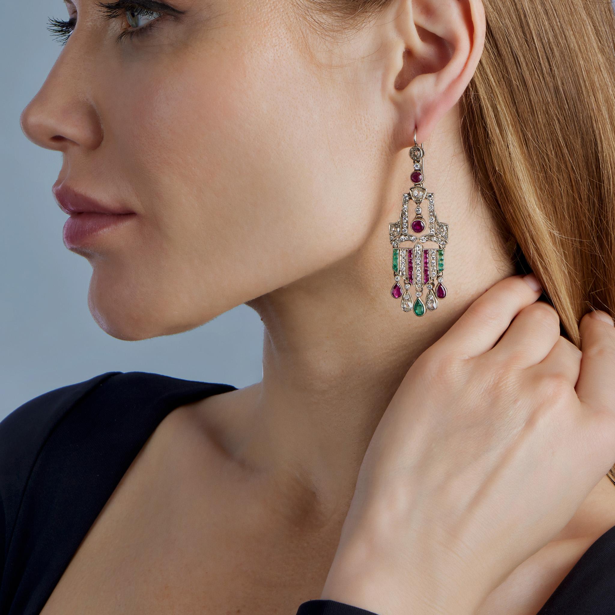 These platinum ruby, emerald and diamond pendant earrings were created circa 1915-1925, in the Late Edwardian to early Art Deco period. Each is designed with a diamond and ruby top suspending a cascade of geometric elements and flexible fringe set
