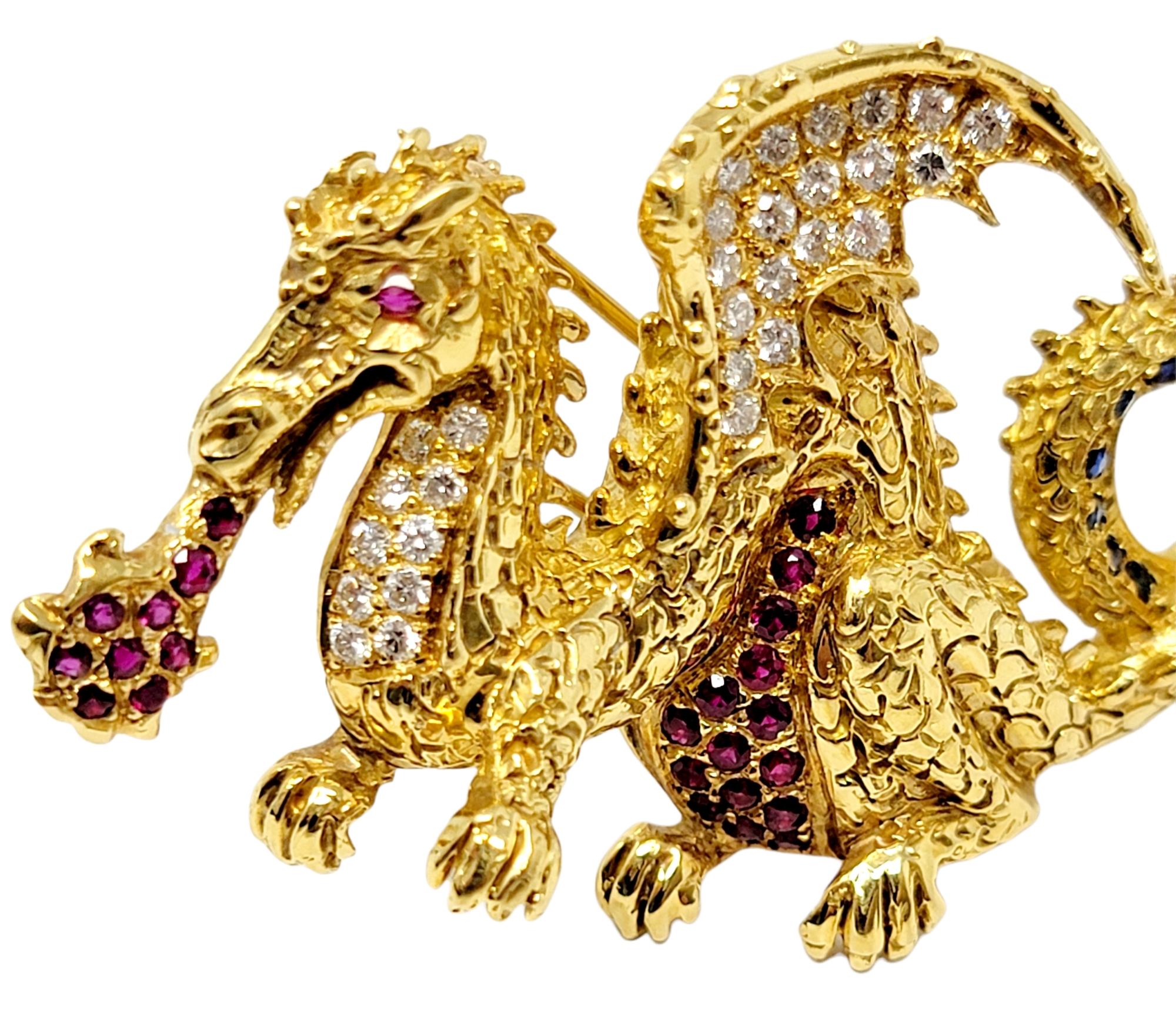 Stunningly detailed fire-breathing dragon brooch accented with glittering diamonds, rubies and sapphires. Made of solid 18 karat yellow gold, the bold dragon has an intricately scaled texture, a looping tail and full wings, which all accentuate the