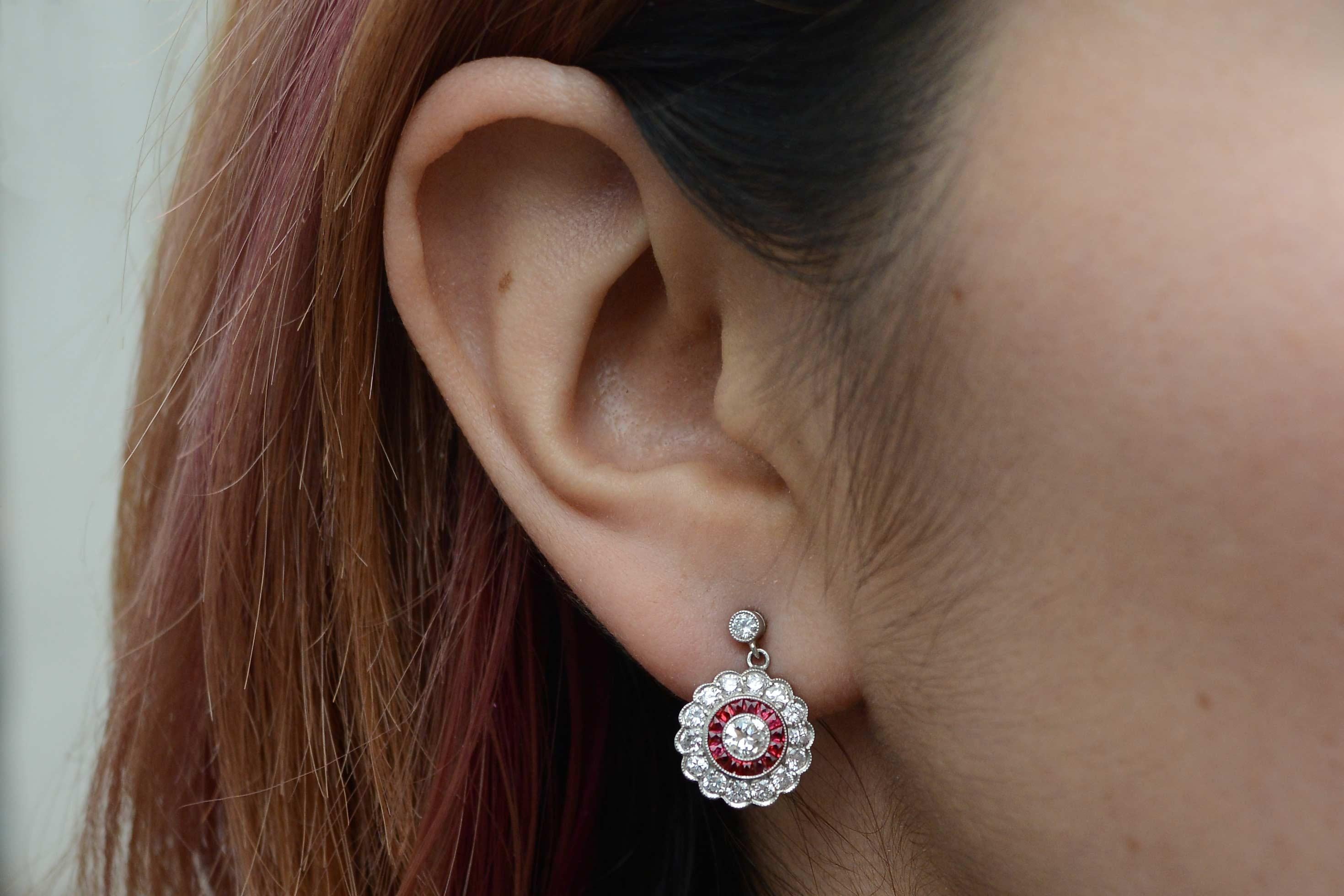 A strikingly rich and decadent combo of fiery diamonds and juicy rubies team up in these Edwardian revival drop earrings. With the two periods often crossing over into the Art Deco era, these scalloped, floral motif halo dangles are formed by a