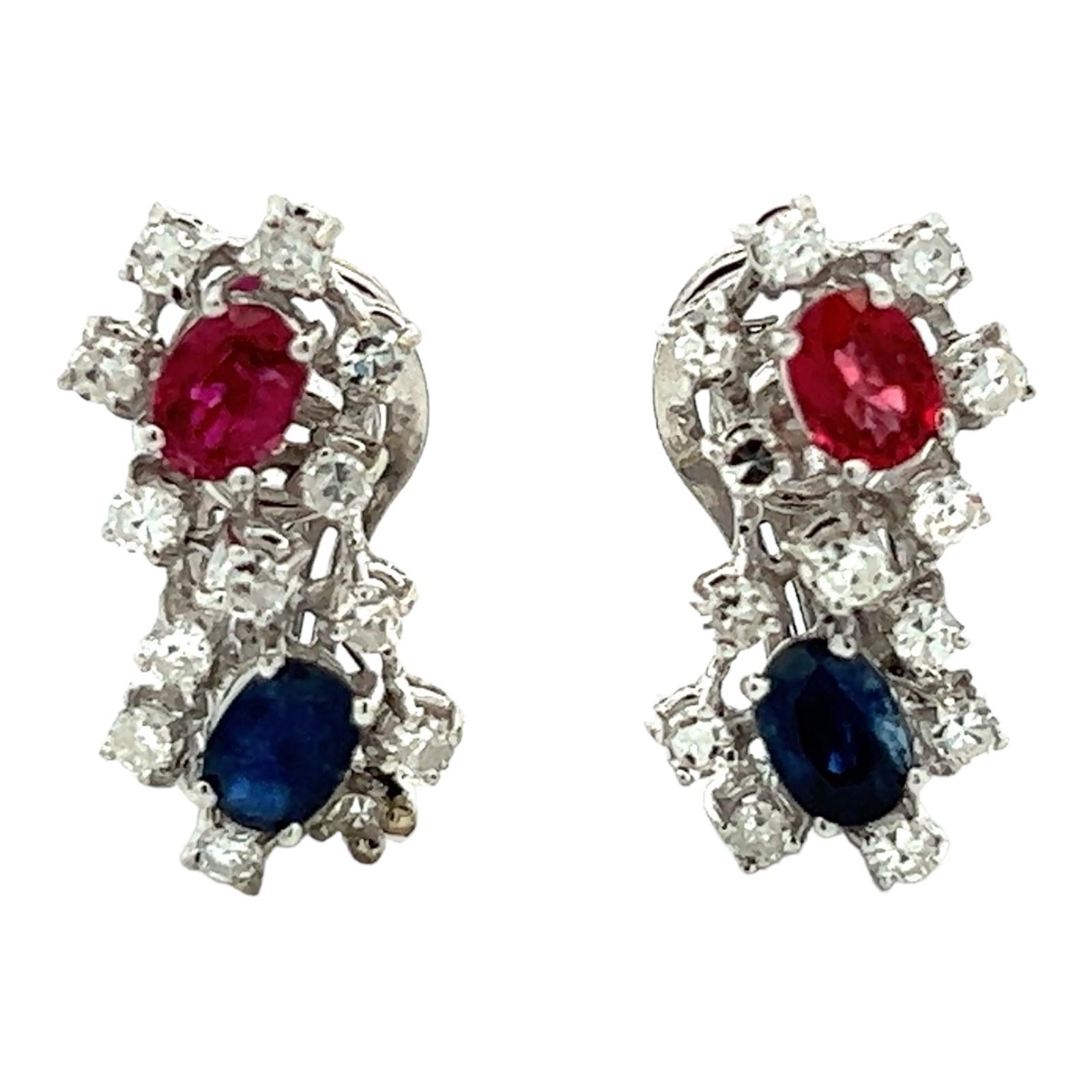 Diamond, sapphire, and ruby drop earrings crafted in 18 karat white gold. The earrings feature 2 oval blue sapphires weighing approximately .80 CTW, and two oval ruby gemstones weighing approximately .90 CTW. The gemstones are surrounded by 20