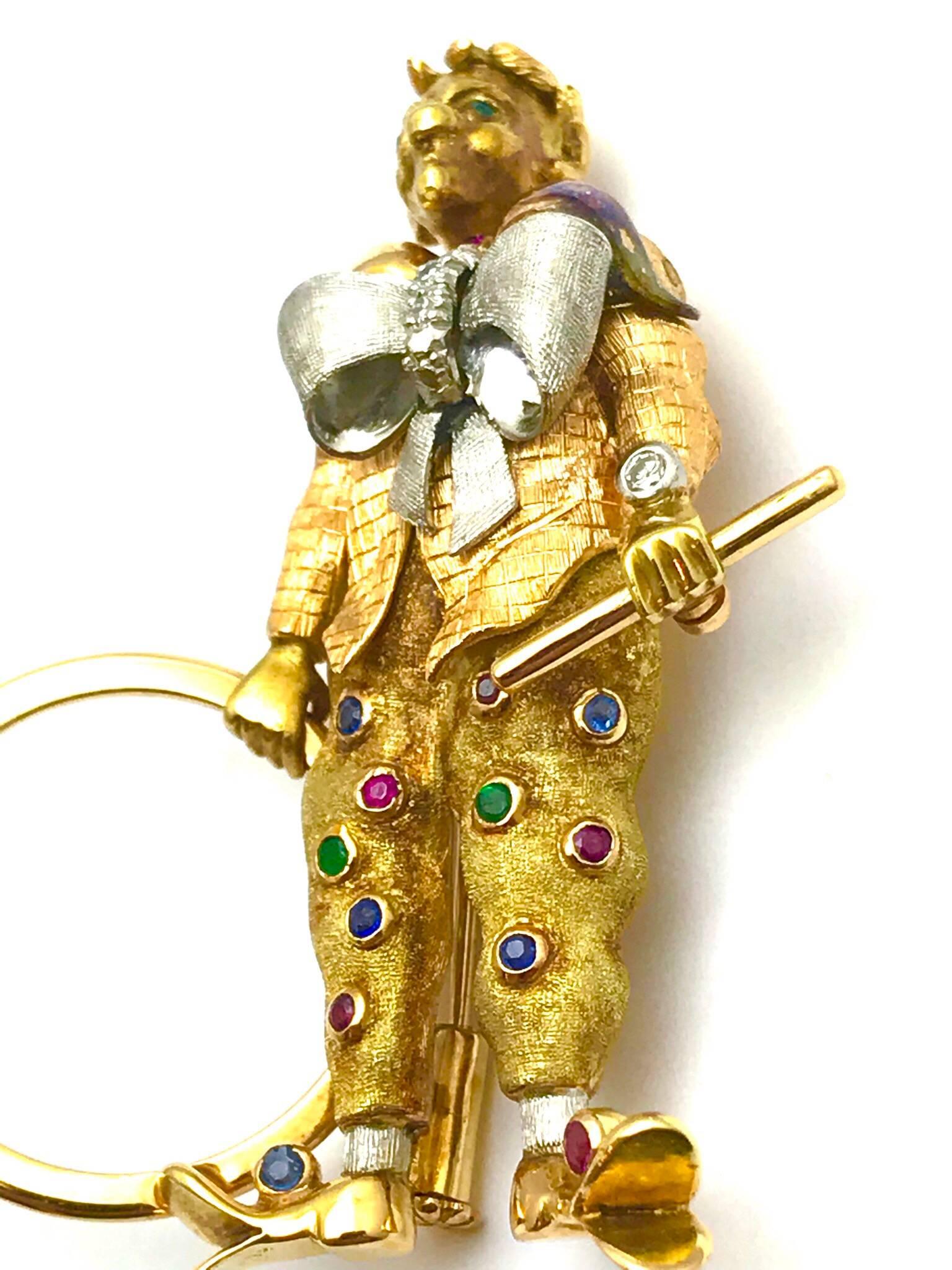 A diamond ruby sapphire and emerald 18 karat rose and white gold circus clown.  He is wearing duck clown shoes, a large white gold bow tie with diamonds, and clown pants sprinkled with ruby sapphire and emeralds, while holding his hoop and staff. 