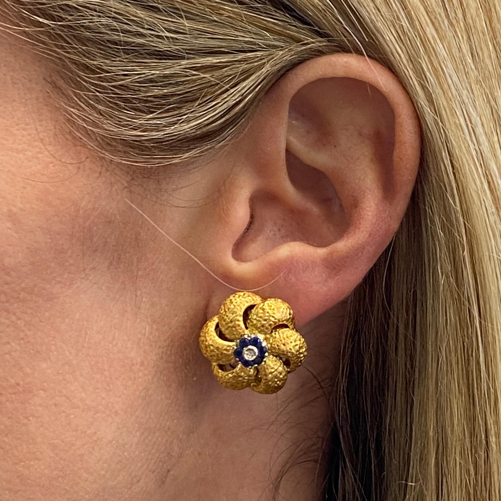 Diamond and sapphire floral earrings fashioned in textured 18 karat yellow gold. The earrings feature 2 diamonds and 12 round blue sapphire accents. The earrings measure 20mm in diameter, and clip backs (posts can be added).

