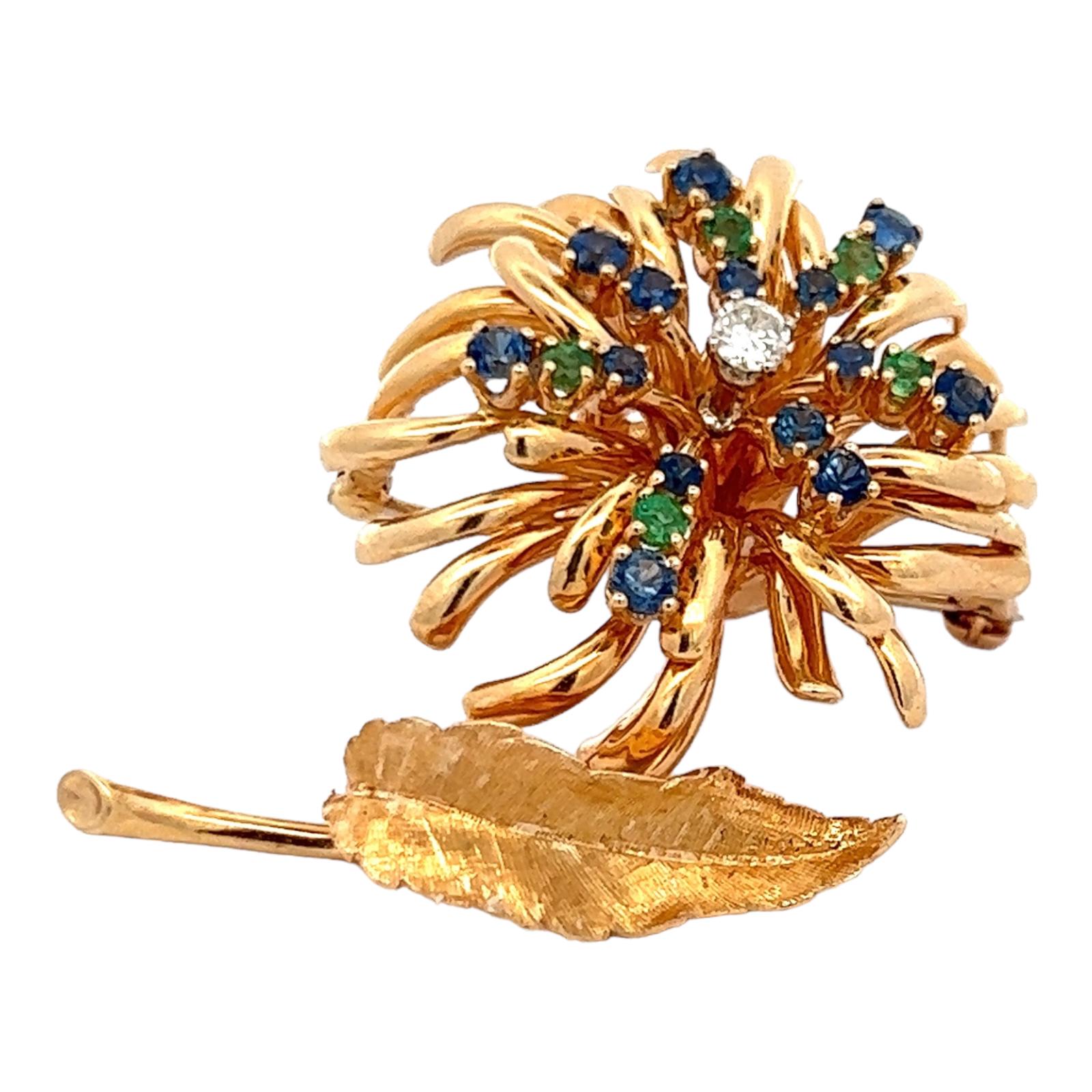 Diamond, sapphire, and emerald floral brooch handcrafted in 14 karat yellow gold. The flower features a center round brilliant cut diamond weighing approximately .15 carat and graded G color and VS clarity. The colorful petals are set with 5 green