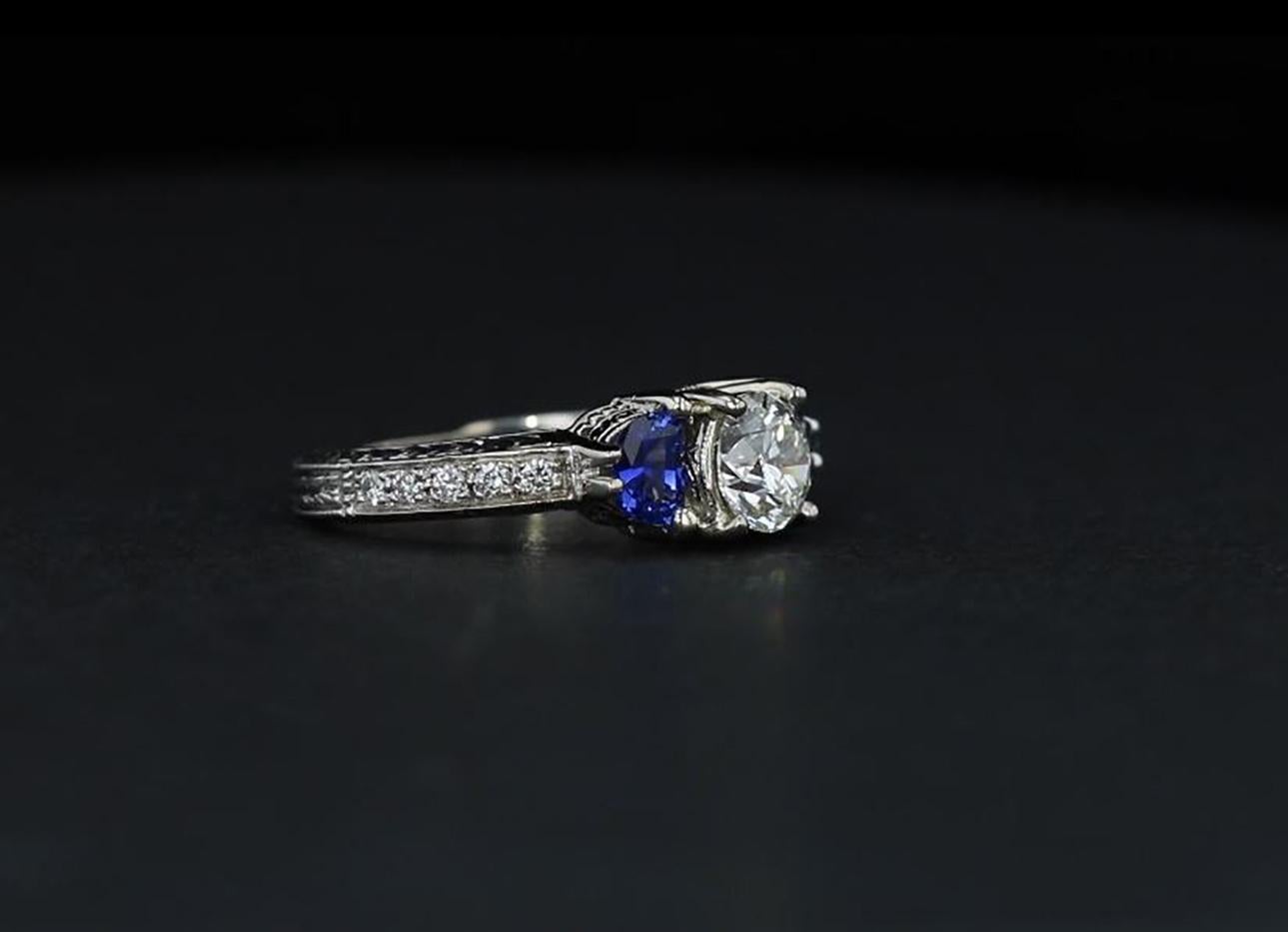 A contemporary take on the classic three-stone engagement ring, a 1.00 carat round diamond in the center of a 14k white gold setting is framed by two half-moon blue sapphires. Delicate engraving and diamonds adorn the ring's shank.