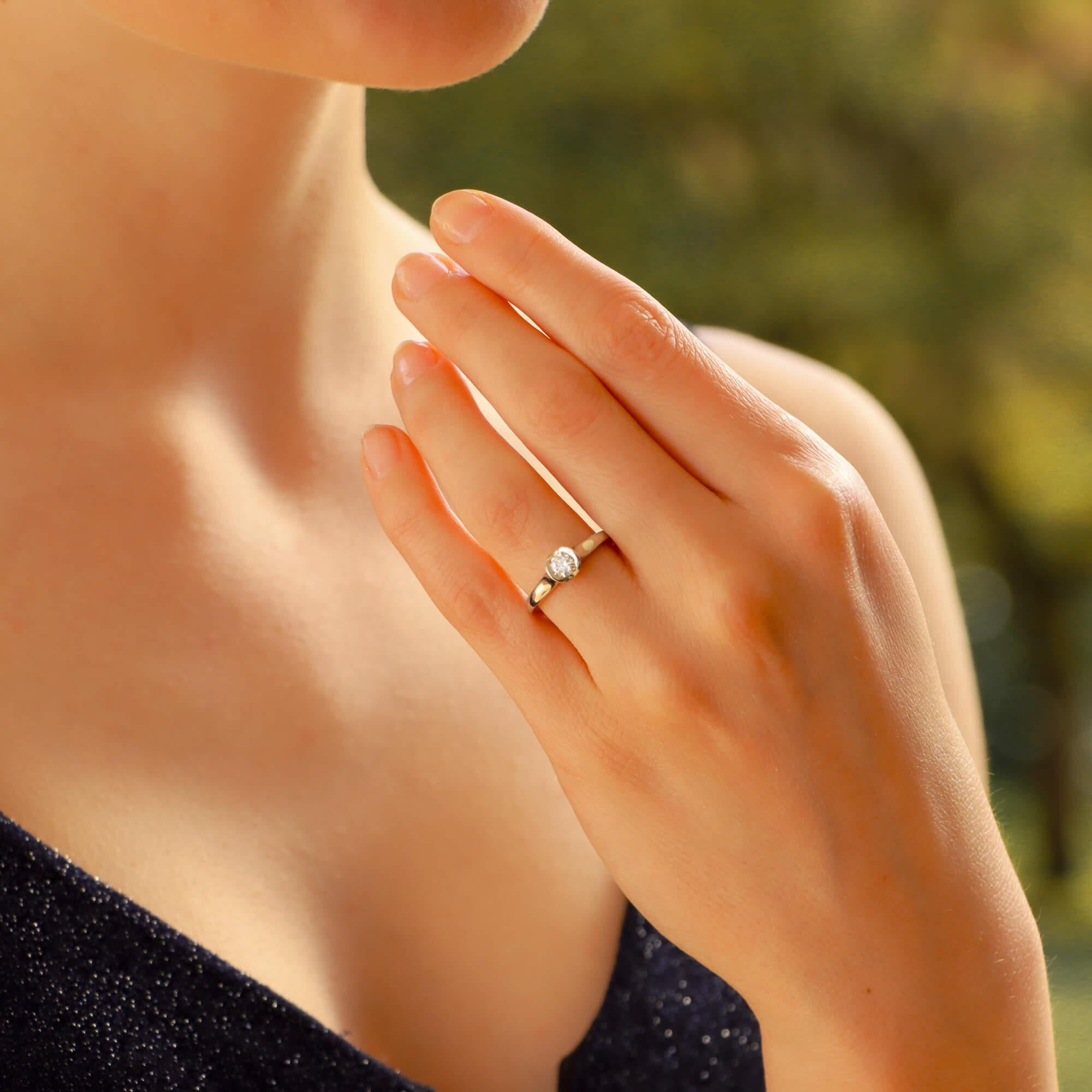 A charming and classic round brilliant-cut diamond solitaire engagement ring in platinum. The ring features a sparkly round brilliant-cut diamond half-rubover-set to the centre in a fancy setting resembling a blooming flower, leading to a solid