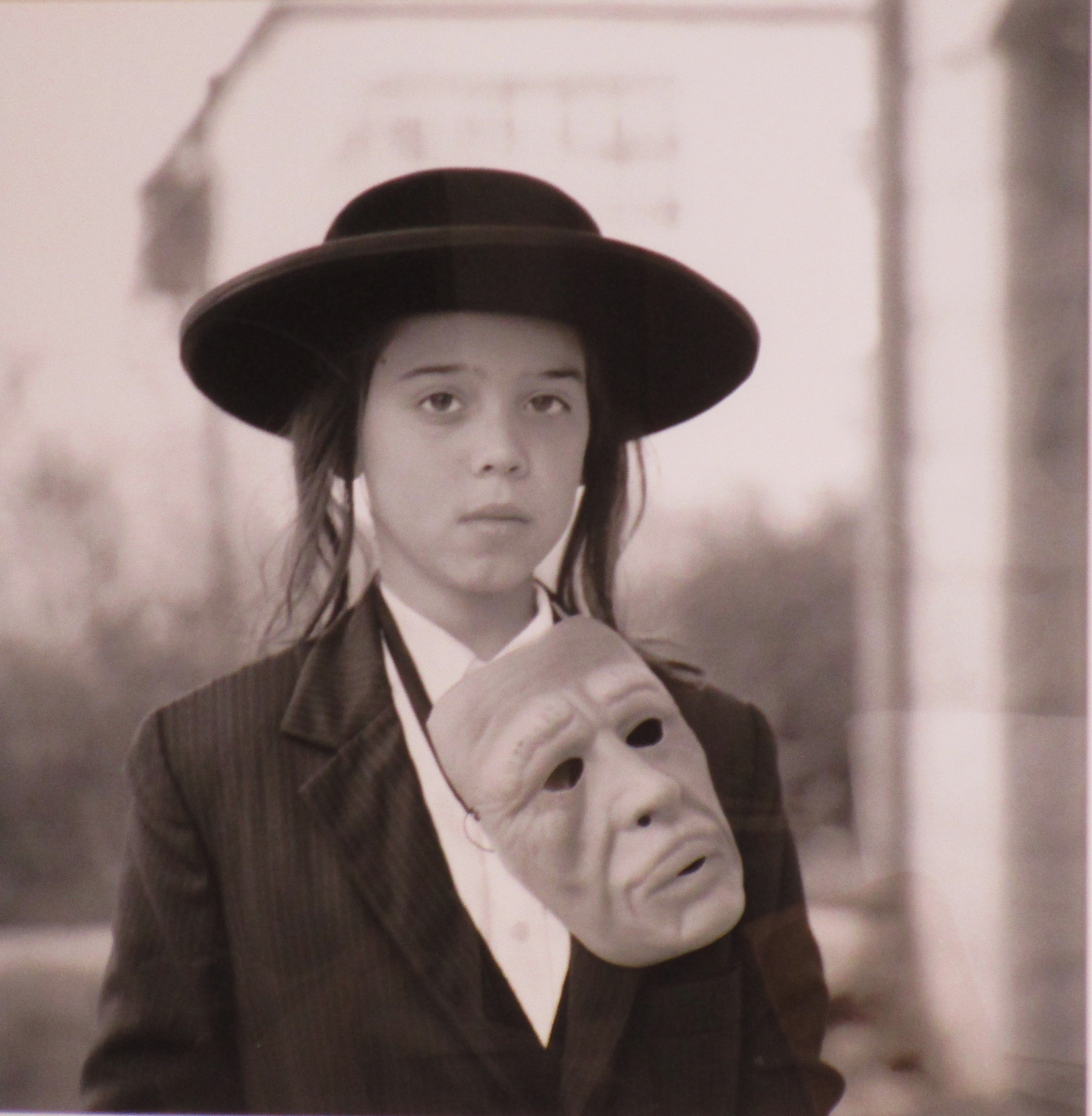 Un hermoso retrato fotográfico en blanco y negro de  joven hasídico con fedora negra y máscara de aspecto inquietante colgada del cuello. Al estilo de Diane Arbus. La foto se enmarcó profesionalmente con un paspartú bajo plexiglás en los años 90. La