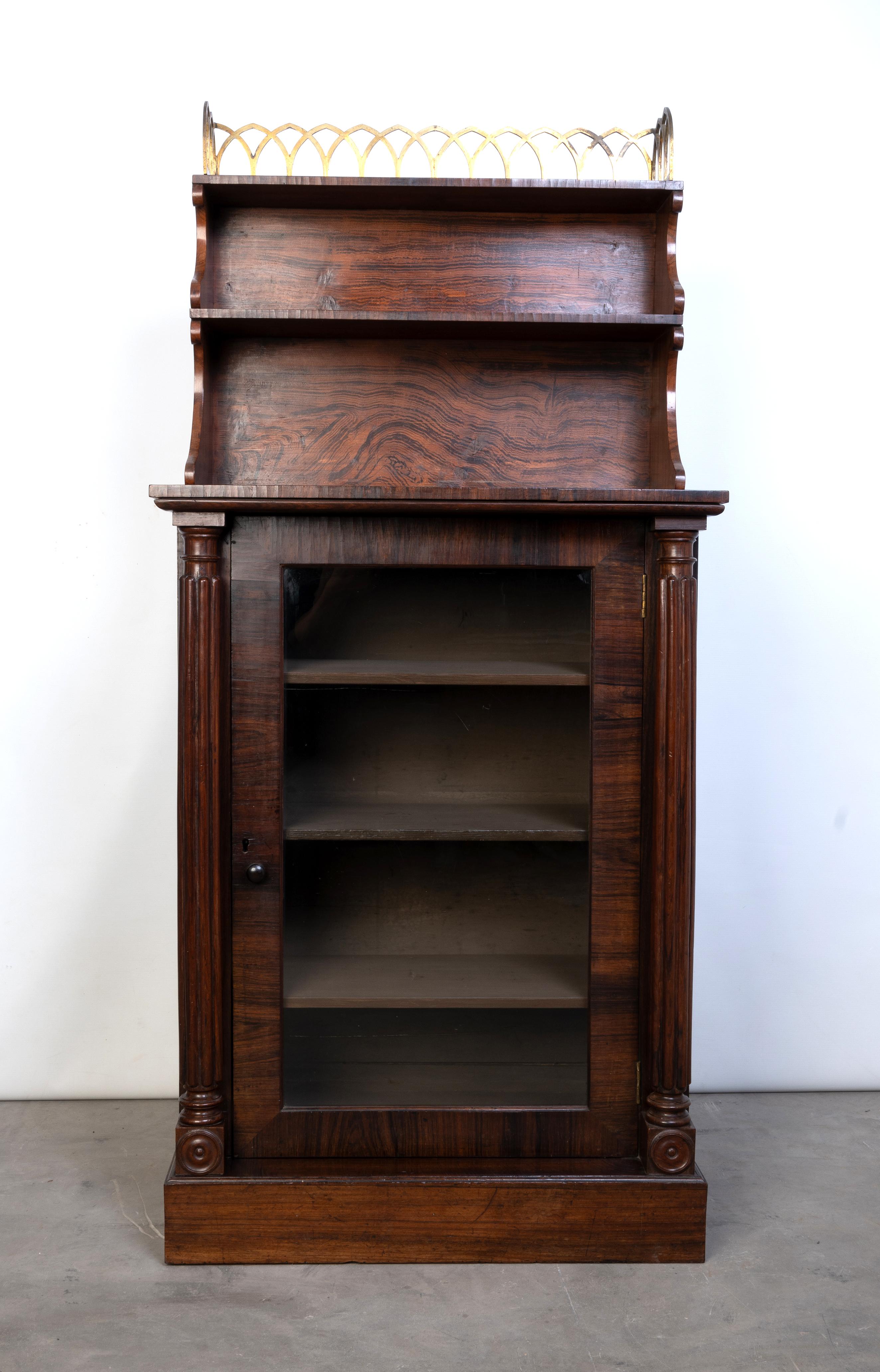 Diminutive Antique English Regency Gilt Brass & Rosewood Side Cabinet C.1820
 
A Regency rosewood cabinet, in the gothic style, with gilt brass gallery and open shelves above a glazed door flanked by gadroon moulded columns, on a plinth base. Of