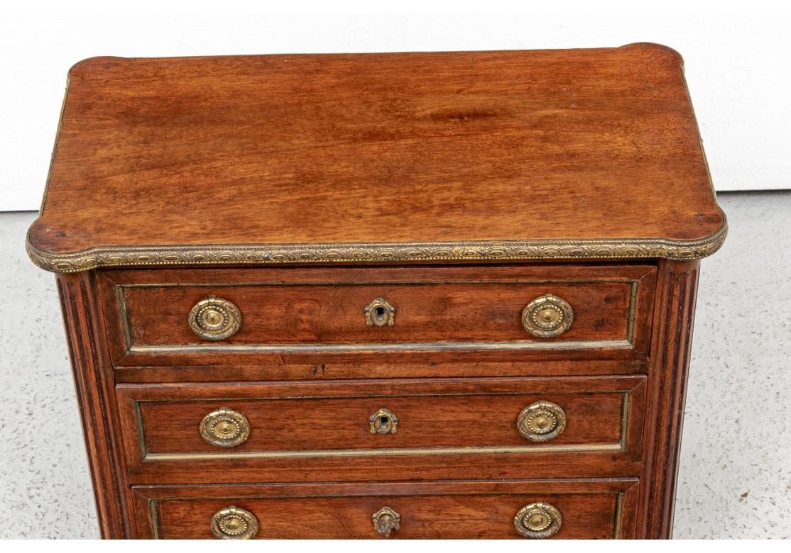 A finely constructed Miniature chest in mahogany with cookie corners on top and banded in brass with ribbon and oval Repoussé motifs. With fluted half column supports front and back raised on turned feet. The three long drawers with brass wreath