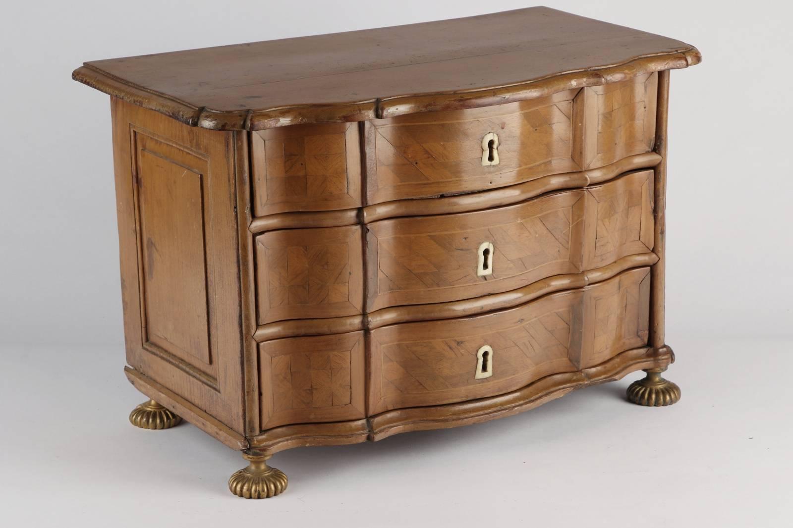 Diminutive fruitwood commode, 18th century, Italian, having three parquetry drawers, bone escutcheons, raised panel sides, scalloped edge and raised on gilt bronze melon form feet.

Condition: Expected wear and signs of use including losses to