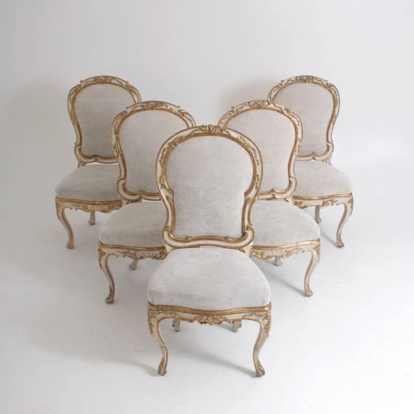 Five dining room chairs on capriole legs with trapezoidal seats and rounded backrest. The chairs are painted in a crème-white color and show gilt ornaments in the shape of c-buckles and bows. Two fitting armchairs are available as well.
   